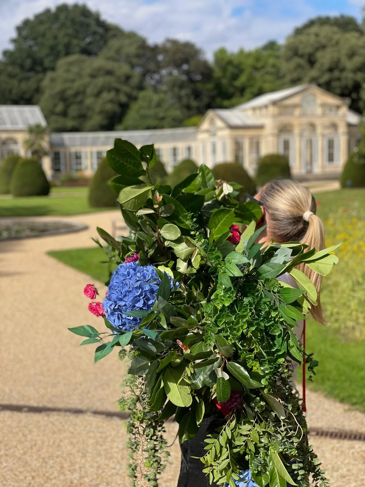 A woman with a ponytail holding a large bouquet of green leaves, pink roses, and a blue hydrangea, standing in front of an old, elegant building with yellow and white trim in a garden setting.