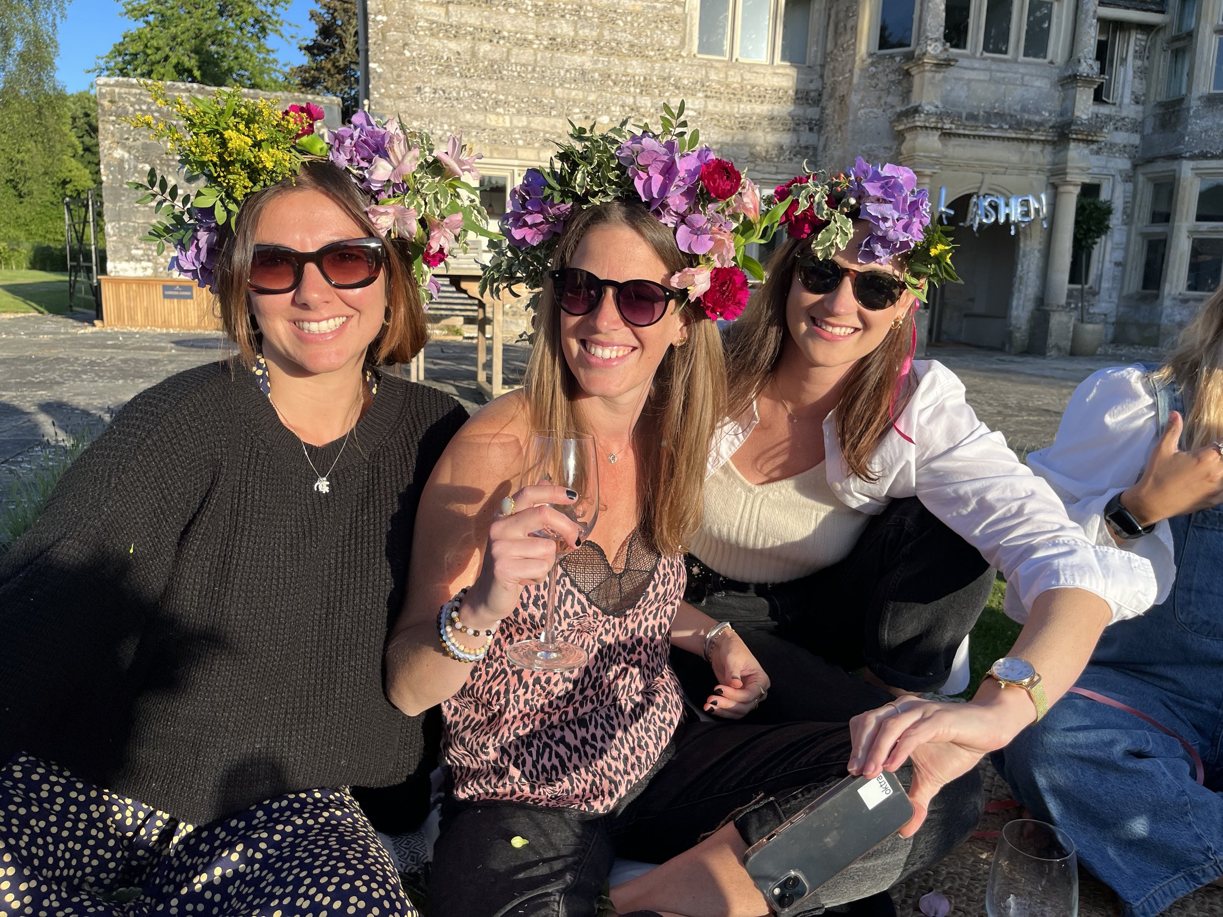 Group of three women wearing flower crowns made of purple, pink, and yellow flowers, sitting outdoors on a sunny day near a large stone building with multiple windows.