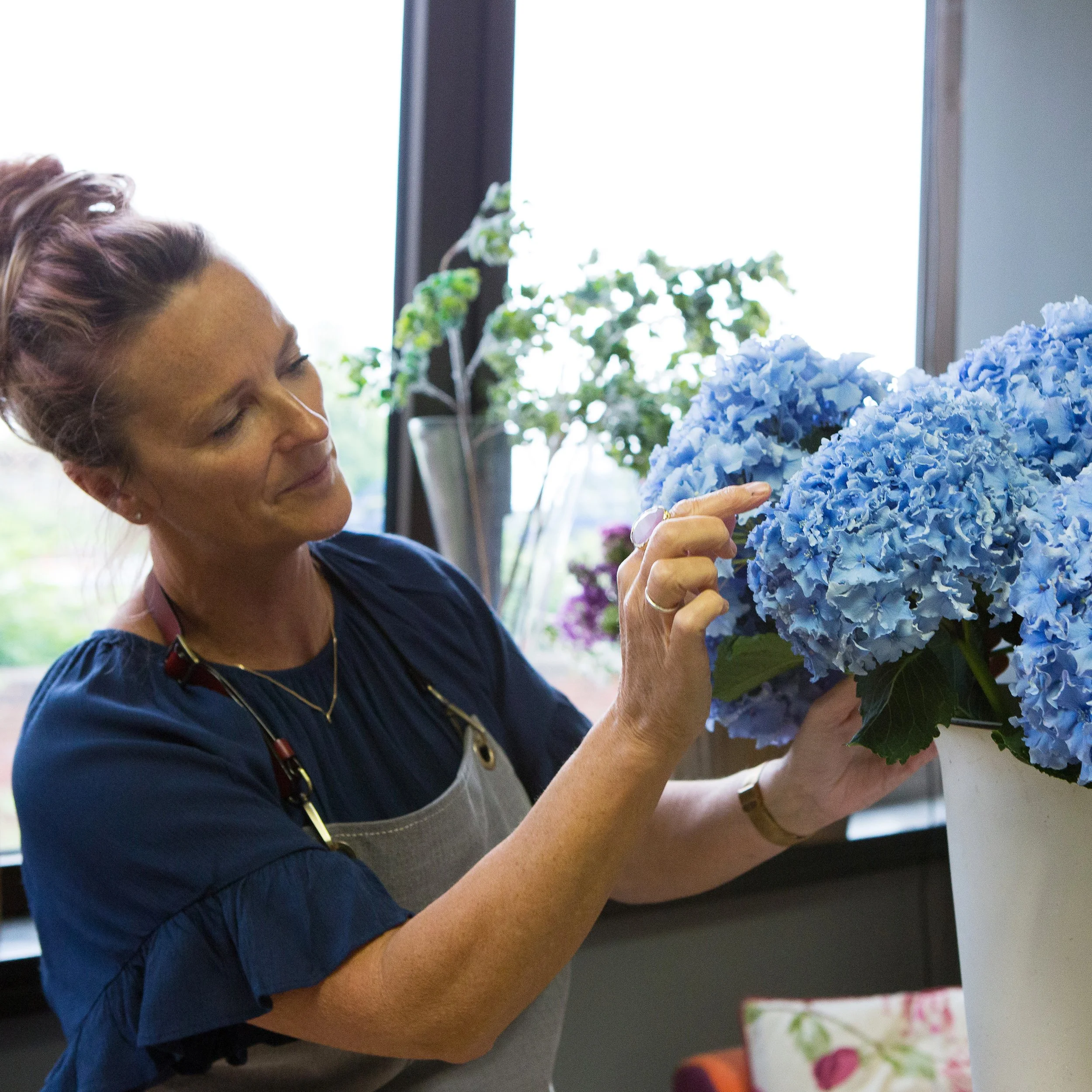 A woman with light brown hair is looking at a blue hydrangea flower cluster on a windowsill, with a tree visible outside through the window.