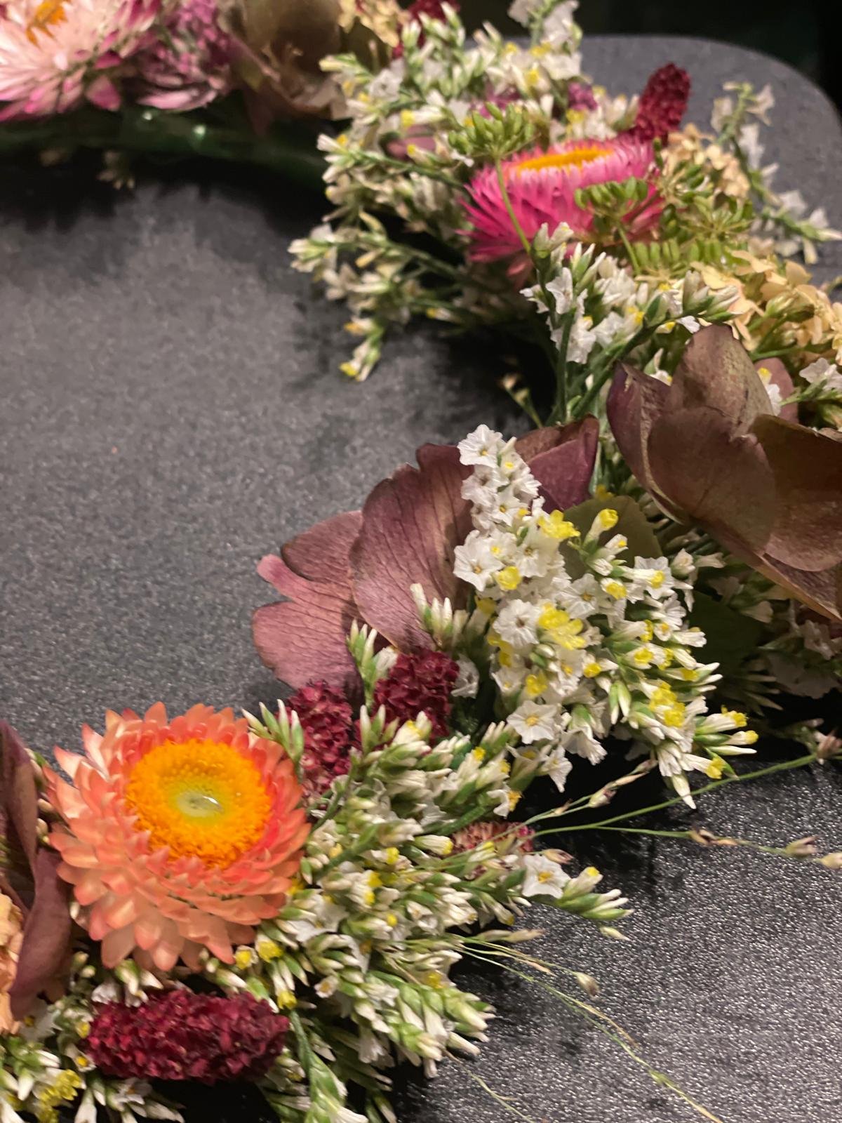 Close-up of a floral wreath made with various colorful flowers including orange, pink, purple, white, and yellow blooms on a dark surface.
