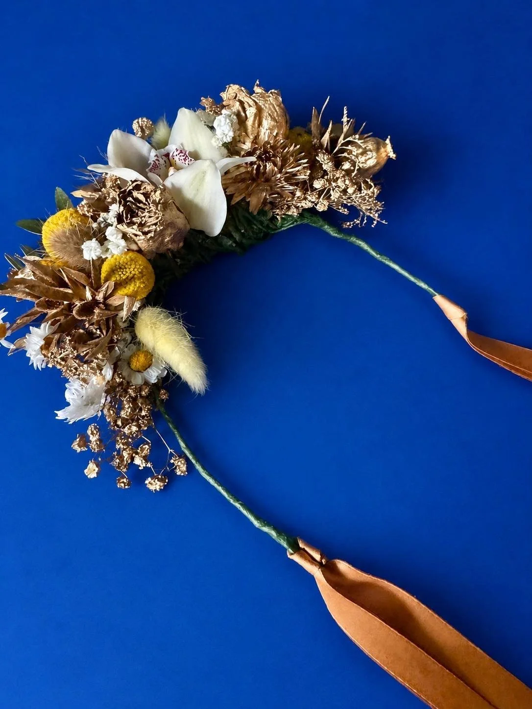 A floral bouquet with dried and fresh flowers, featuring white, yellow, and brown tones, tied to a copper-colored ribbon, against a blue background.