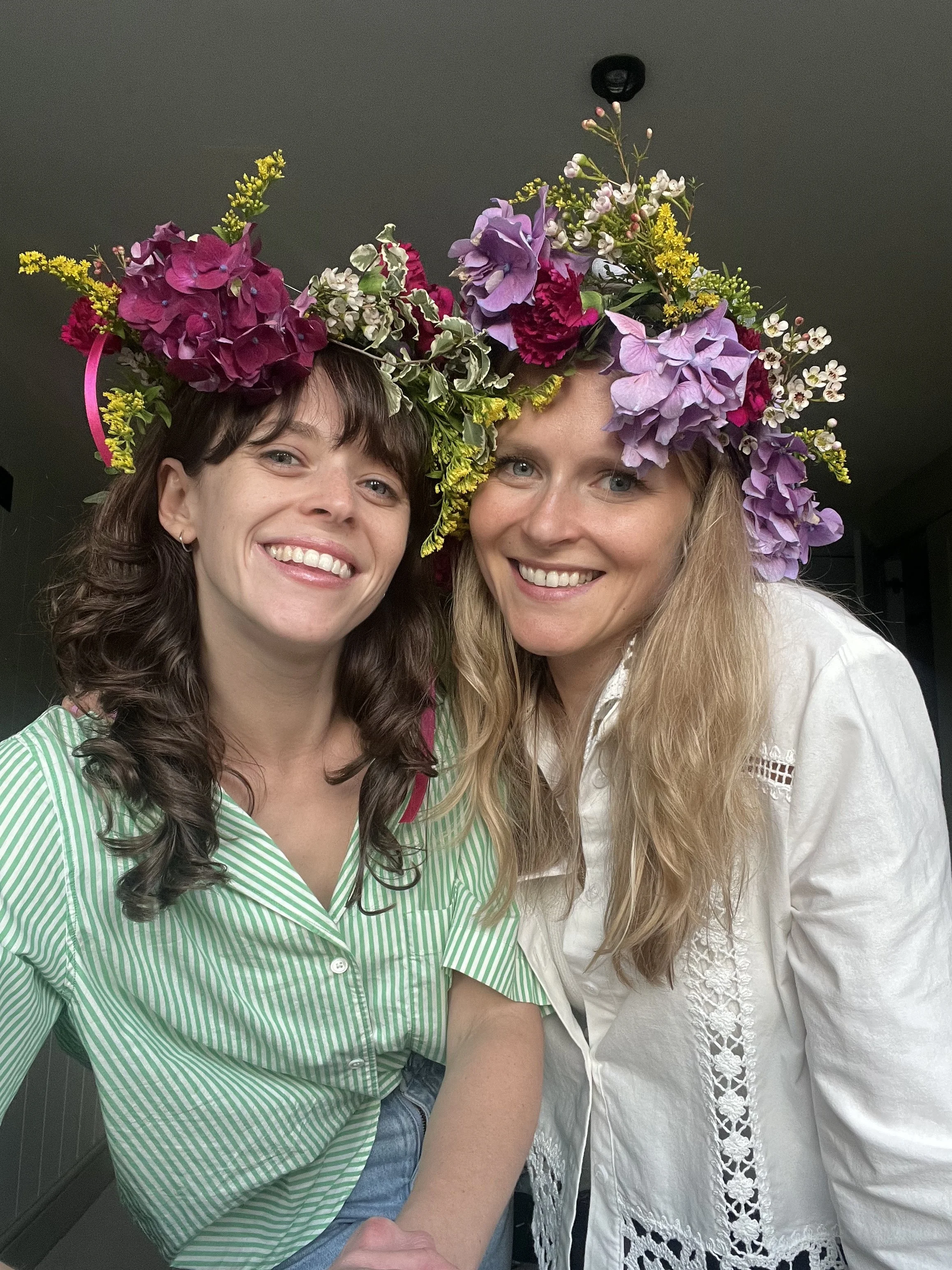 Two women wearing floral crowns smiling for a selfie indoors.