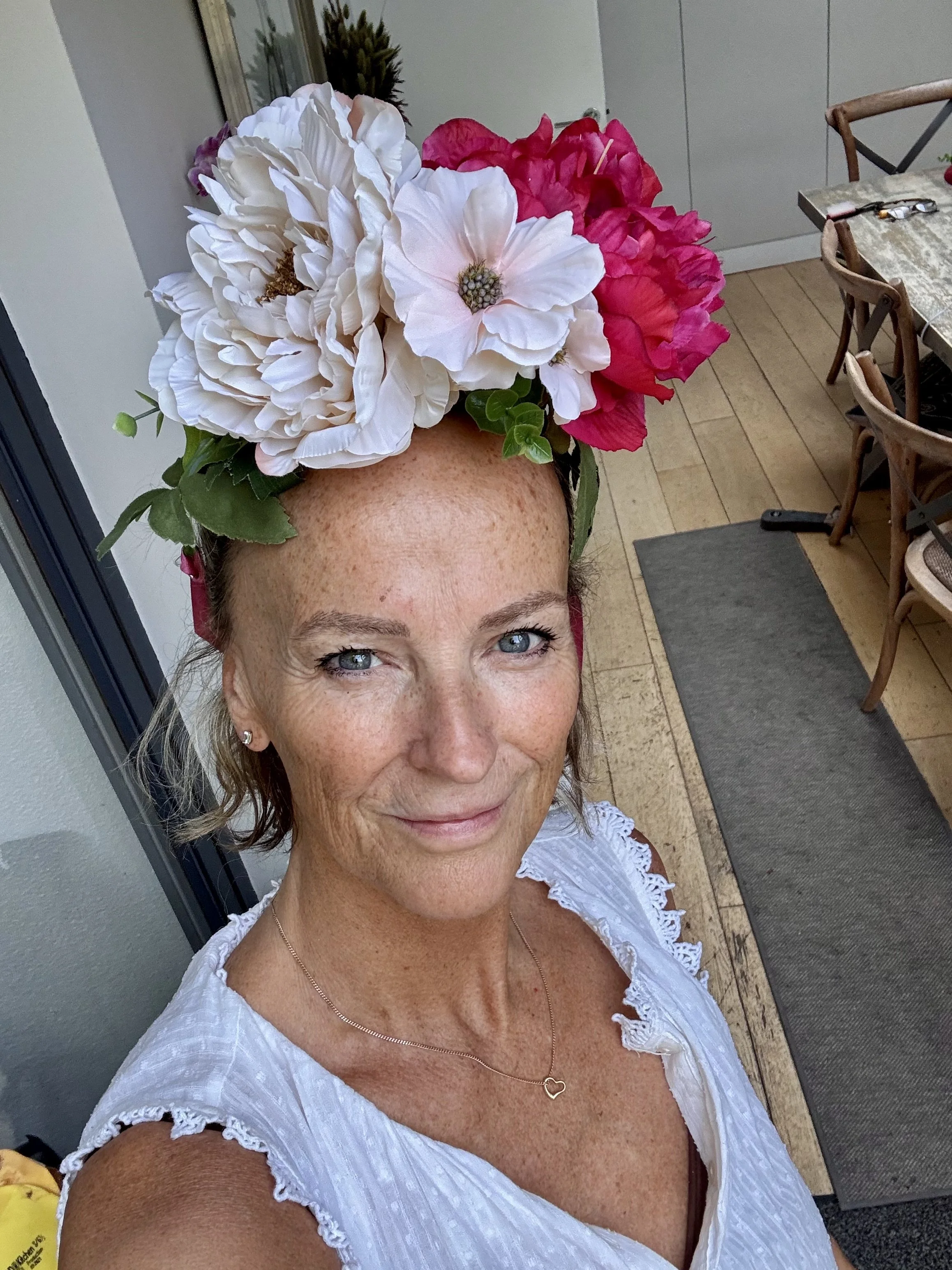 A woman with blue eyes, freckles, and wavy brown hair styled in a side part, wearing a white blouse with lace details and a delicate heart necklace, holding a bouquet of large white and pink flowers on her head, standing outdoors near a patio area wi