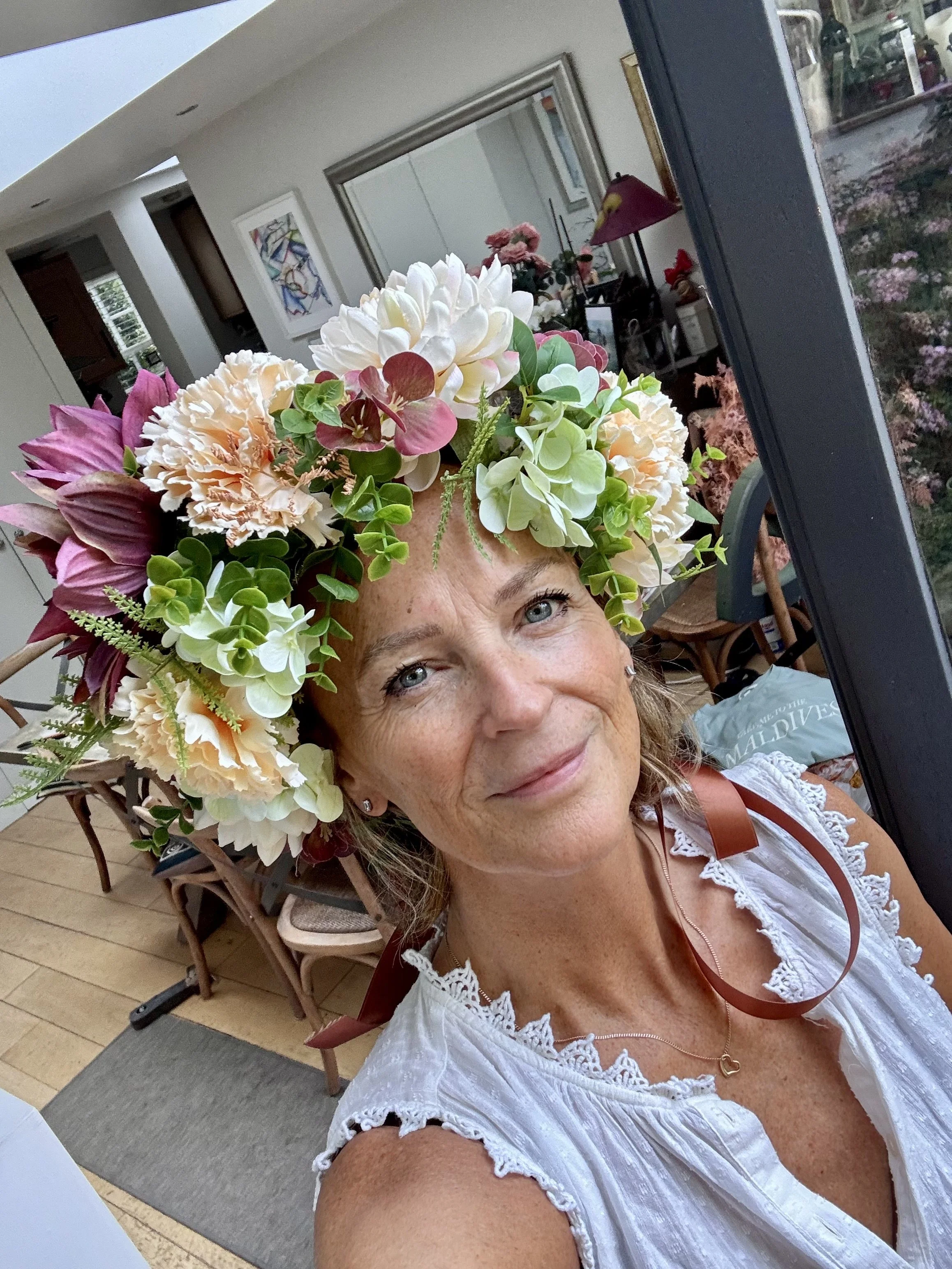 A woman with gray eyes and a nose piercing taking a selfie indoors, wearing a floral crown made of various flowers and greenery, and a white eyelet top with lace trim.