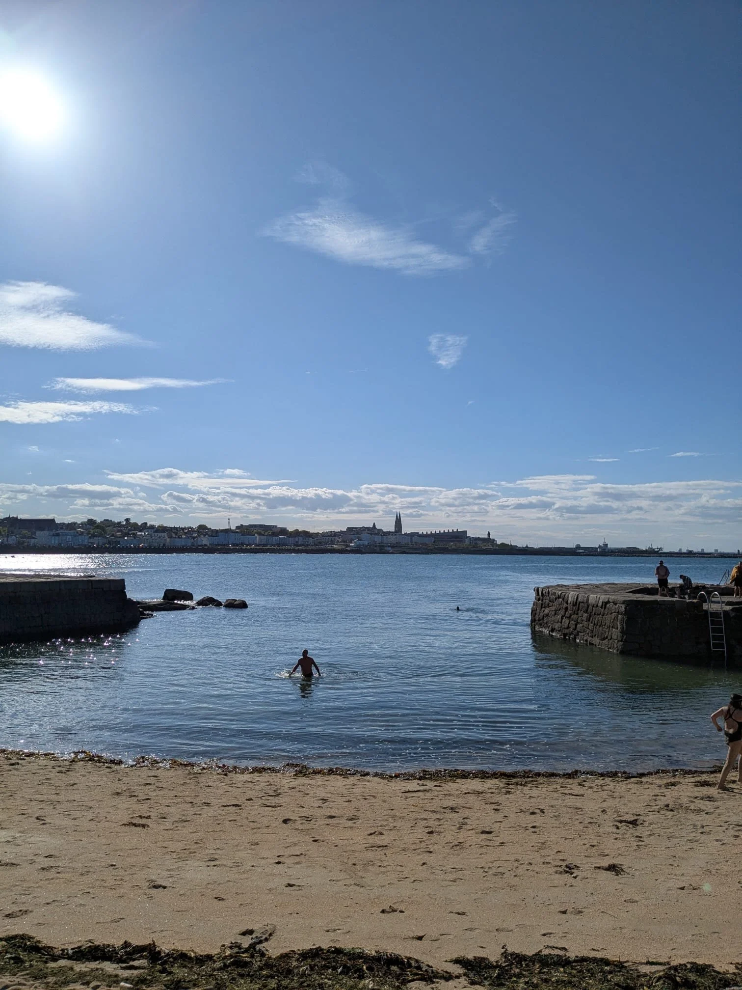 A beach scene with a sandy shore in the foreground, a person swimming in the water, and a pier with stone supports extending into the water. The background shows a city skyline with various buildings, including a tall church steeple, under a partly cloudy sky with the sun shining.