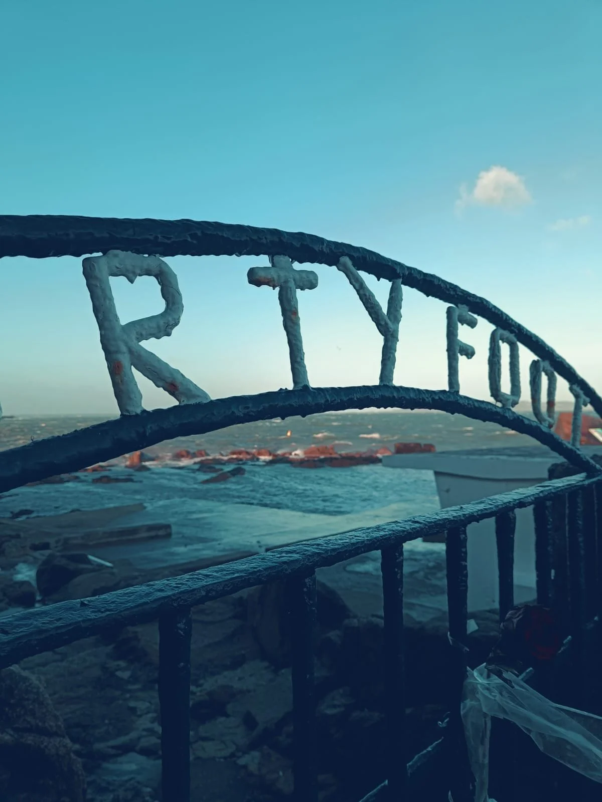 Ice-covered metal railing with the word "RATYEN" on top, overlooking a body of water under a blue sky with a small cloud.