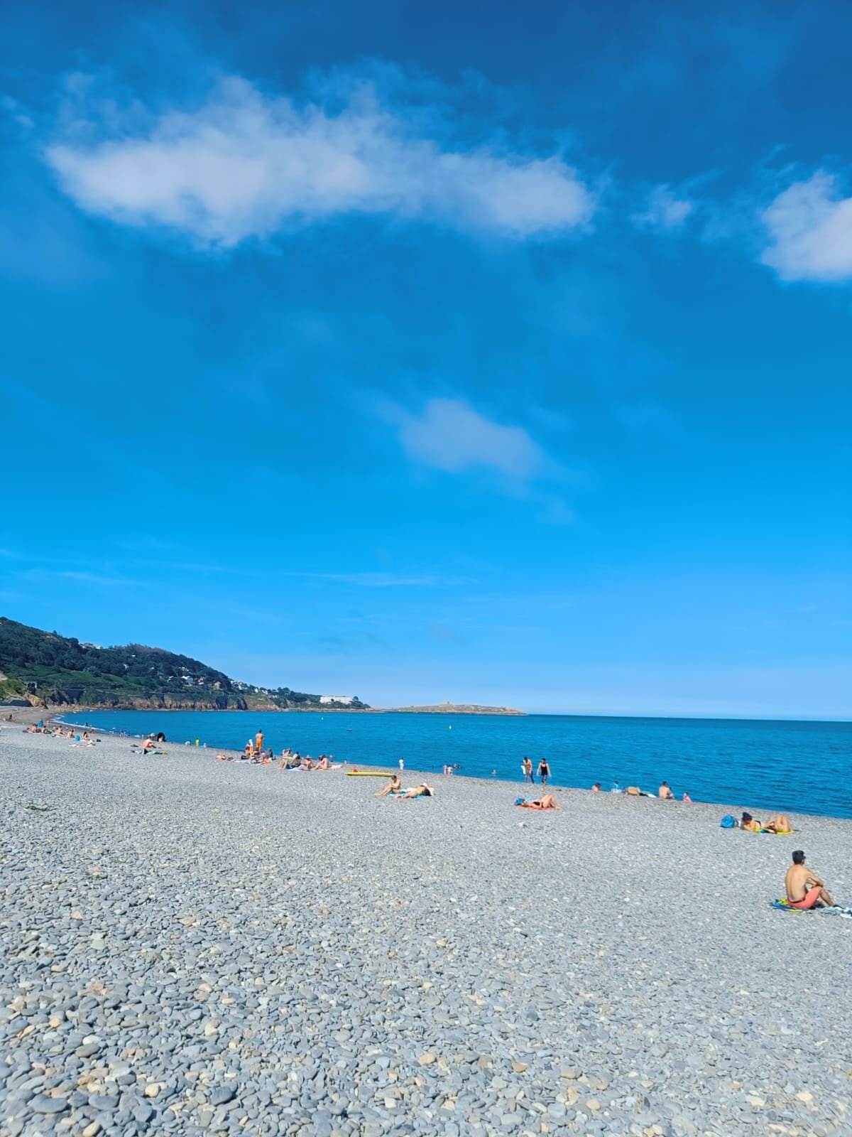 Beach with people sunbathing and swimming under a blue sky with clouds, with a hilly coastline in the background.