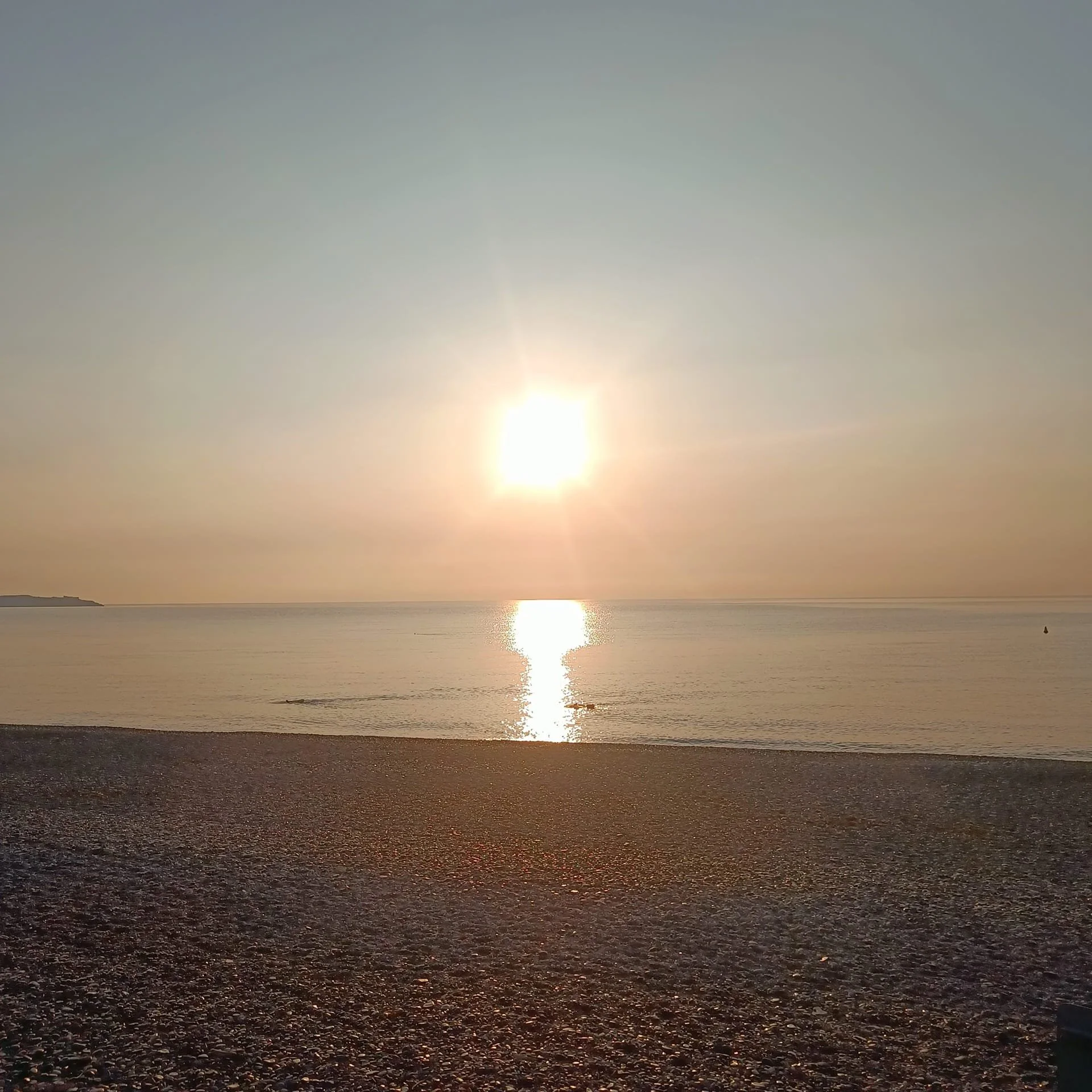 Sunset over calm ocean with a pebbled beach in foreground and small silhouette of a boat on the water.
