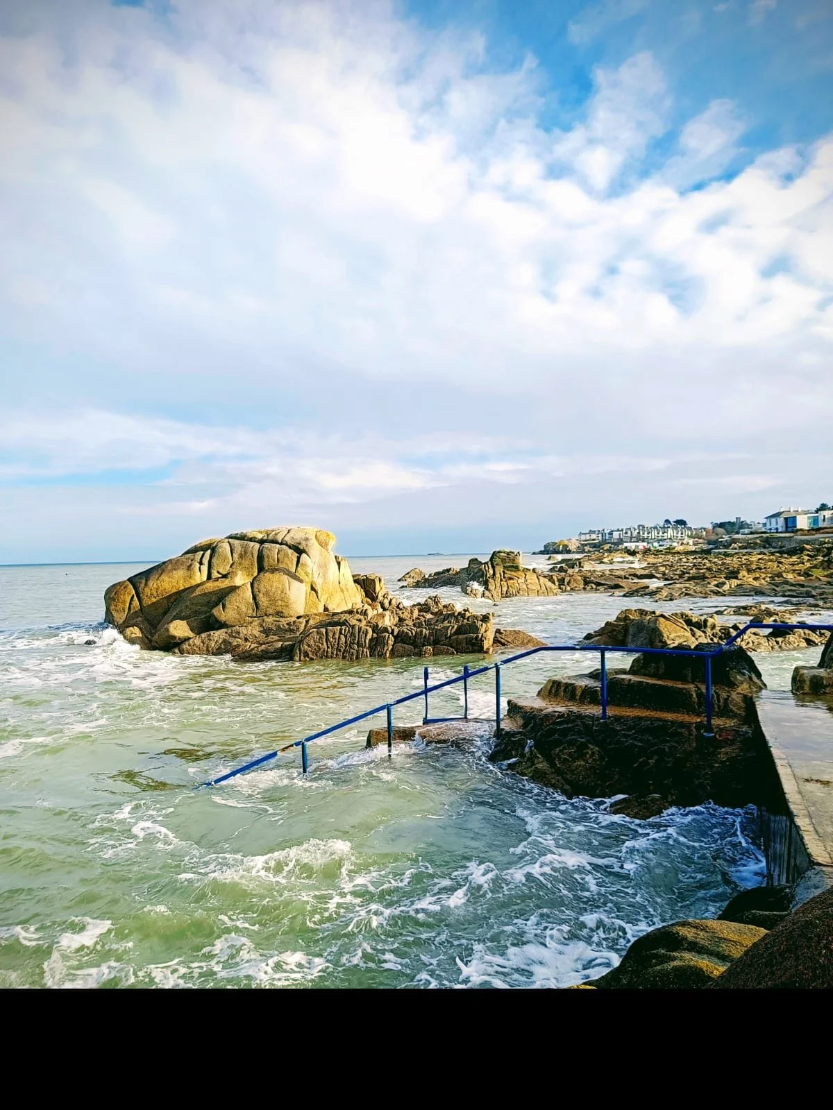 Coastal scene with large rocks by the ocean, a shoreline with buildings in the distance, partly cloudy sky, and waves crashing against rocks with a blue railing along the rocks.