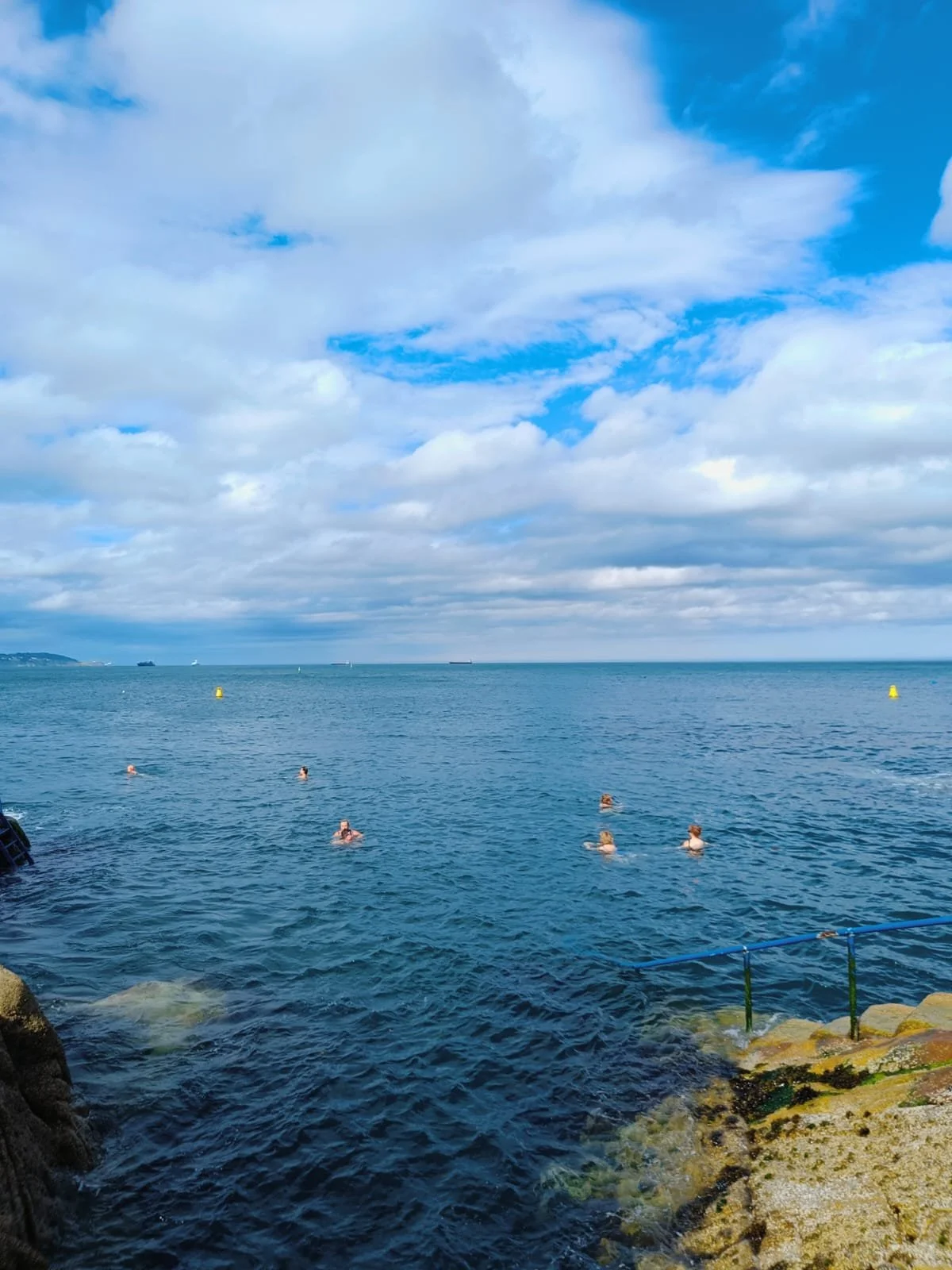 People swimming in the ocean near rocky shore with a cloudy blue sky and ships on the horizon.