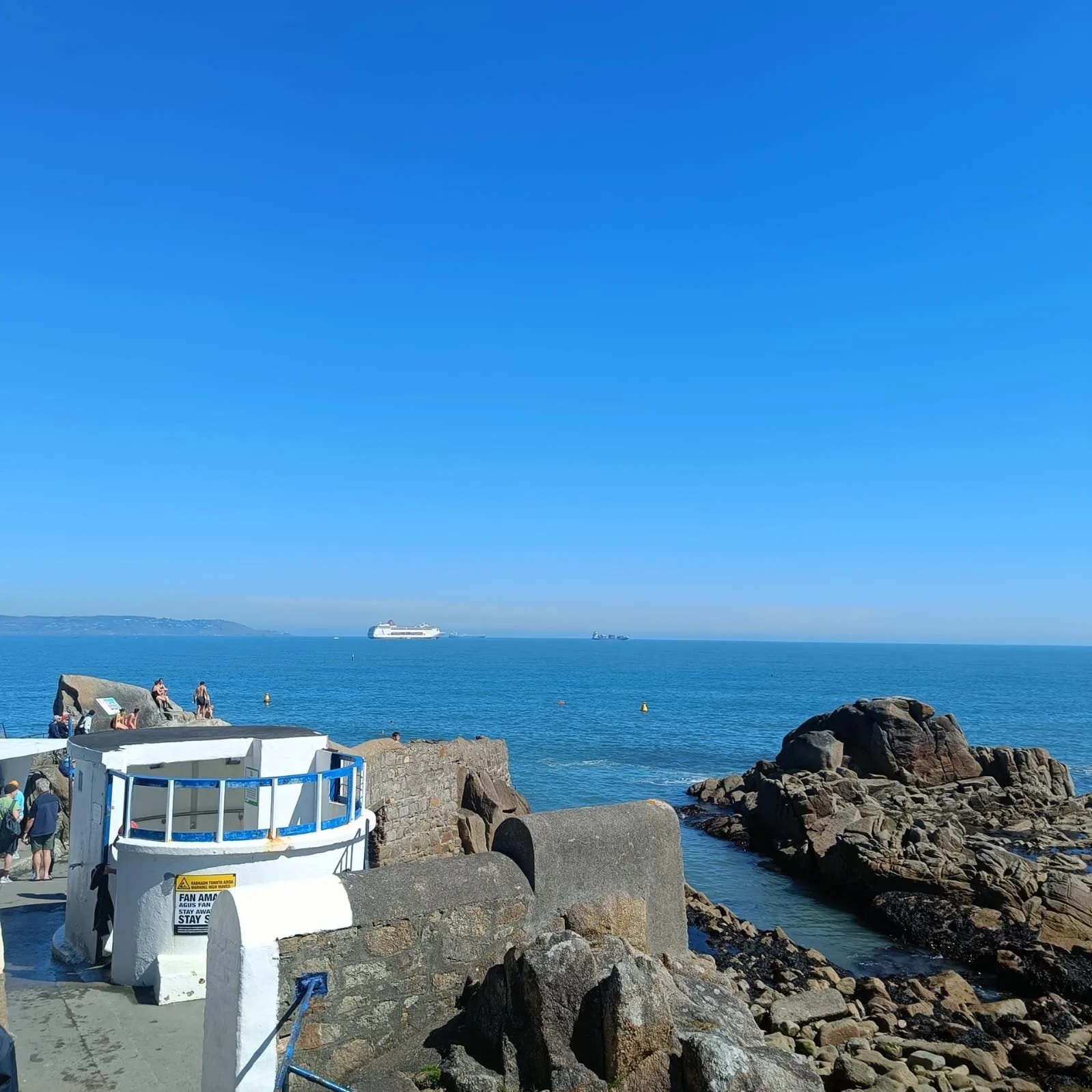 A coastal scene with large rocks and a stone pier in the foreground, in the distance a white cruise ship sails on calm blue water under a clear blue sky, with a few people on the pier.