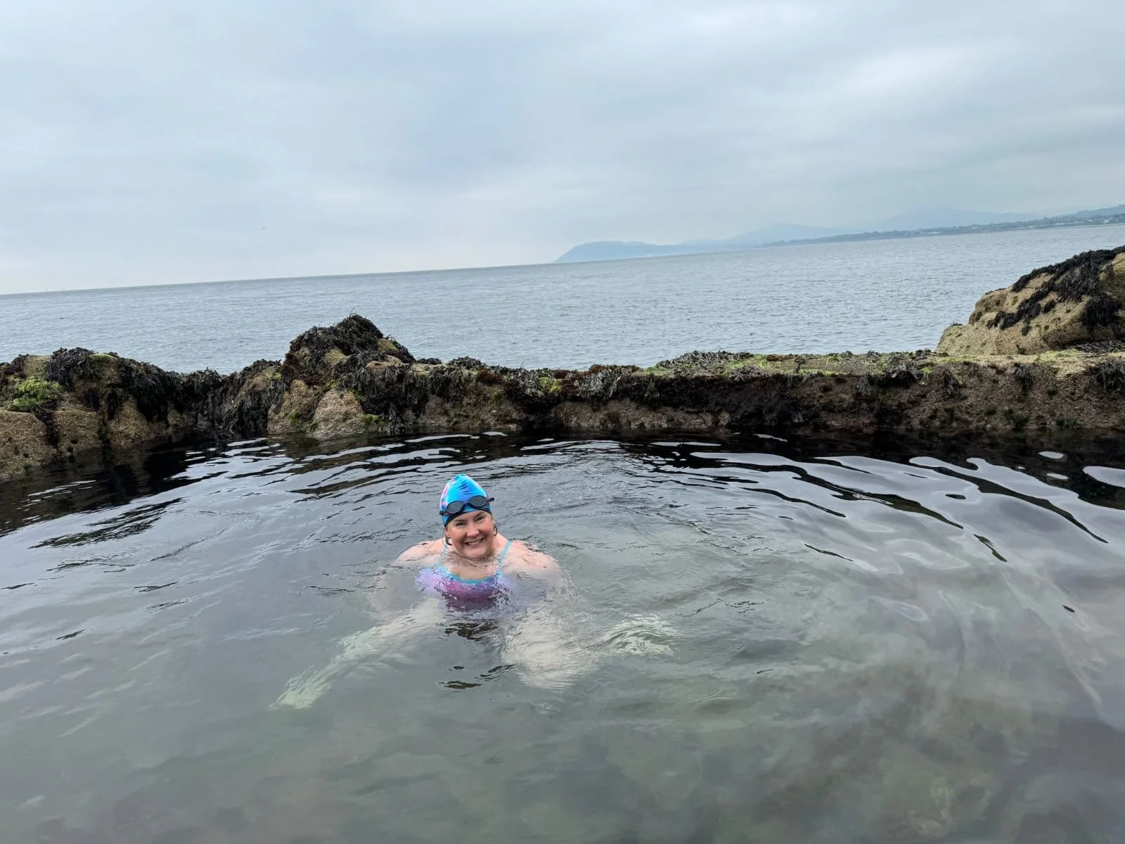 A woman smiling in a swimming pool with rocky edges, wearing a swim cap and goggles, overlooking a body of water on a cloudy day.