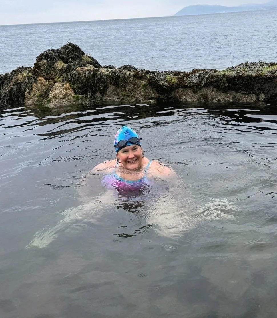 A woman swimming in an ocean or sea, wearing goggles and a blue swim cap, smiling at the camera, near rocks with a distant view of land or mountains.