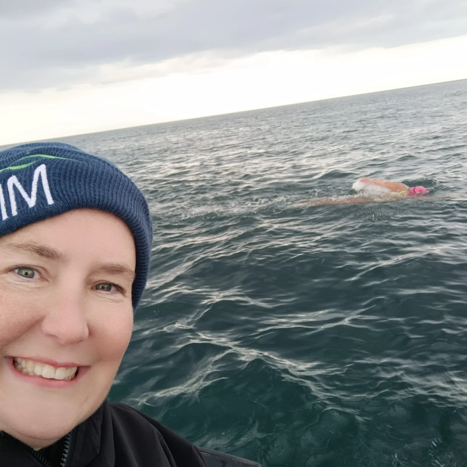 A person smiling and wearing a dark jacket and a blue beanie with the Alaska Airlines logo, with a body of water and a swimmer in a pink swimsuit and pink swim cap in the background.