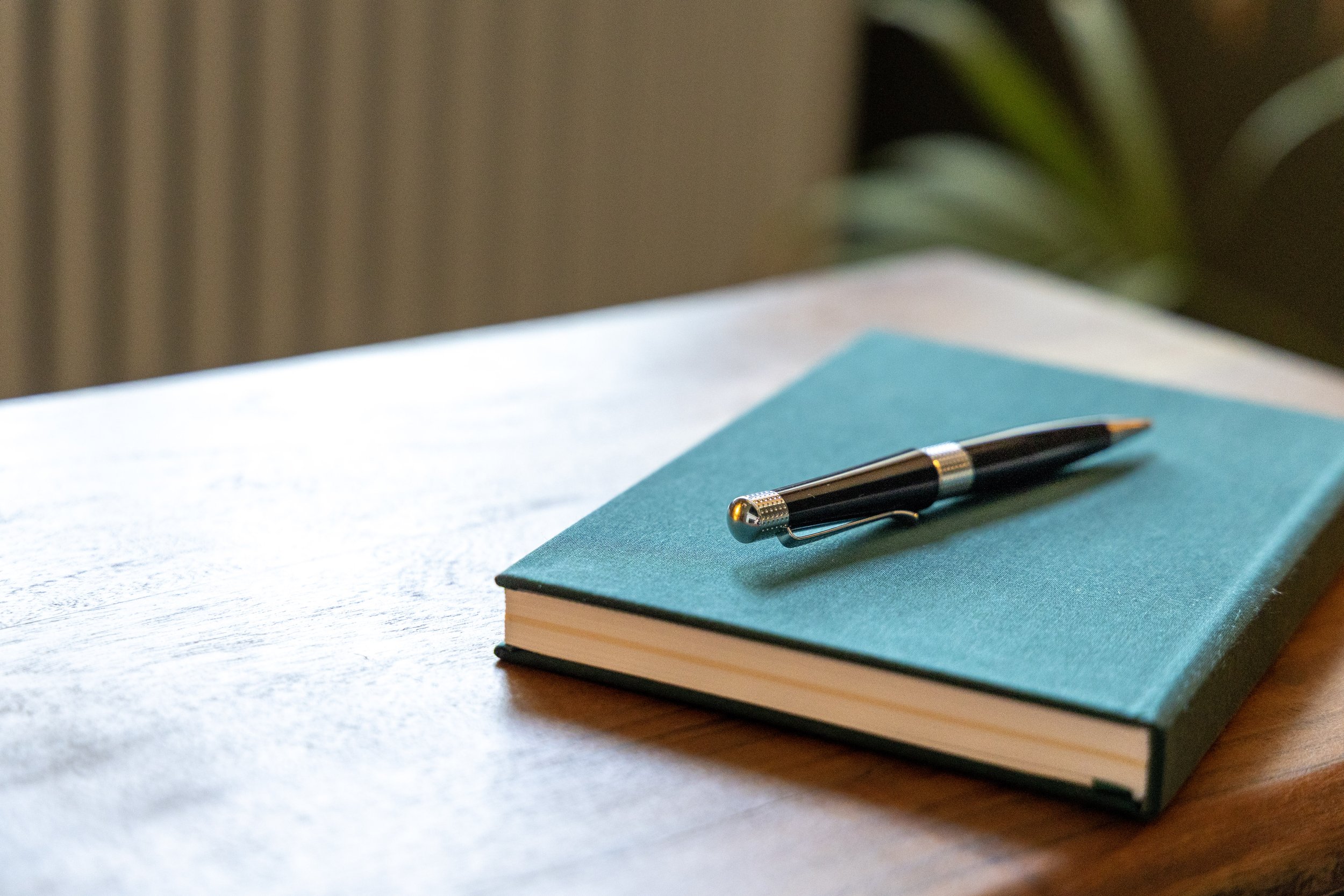 A black and silver pen resting on a closed teal notebook with a black spine, placed on a light-colored wooden table.