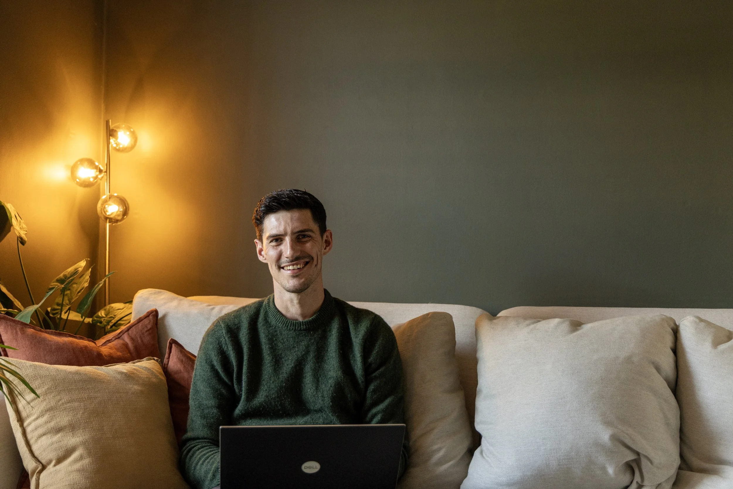 A man sitting on a beige couch with multiple pillows, using a laptop with a Dell logo, in a room with a yellow wall light and green plants.