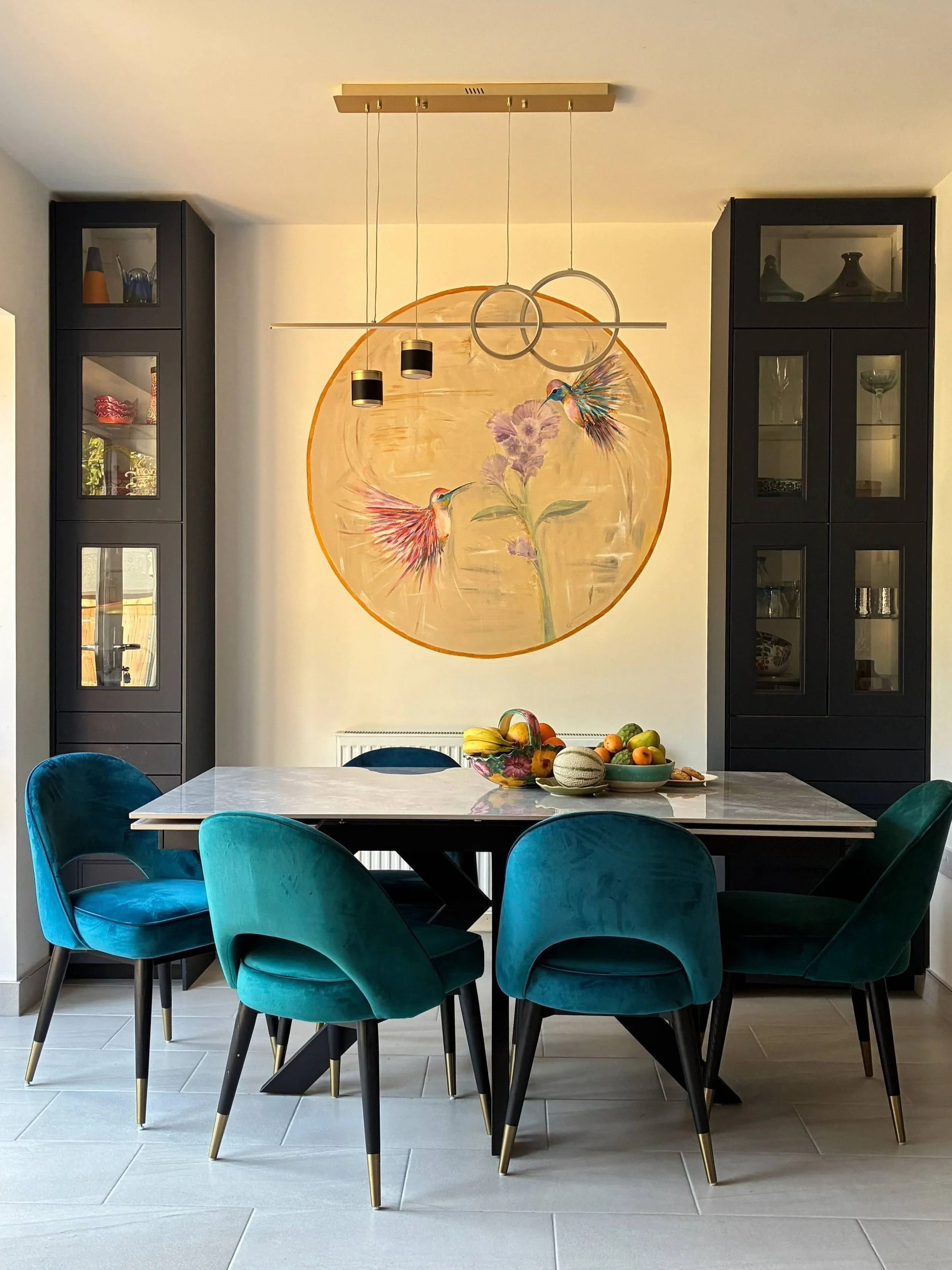 Modern dining room with a white marble table, six teal velvet chairs, black cabinets with glass doors, a fruit bowl, floral wall art with hummingbirds, and gray tiled floor.