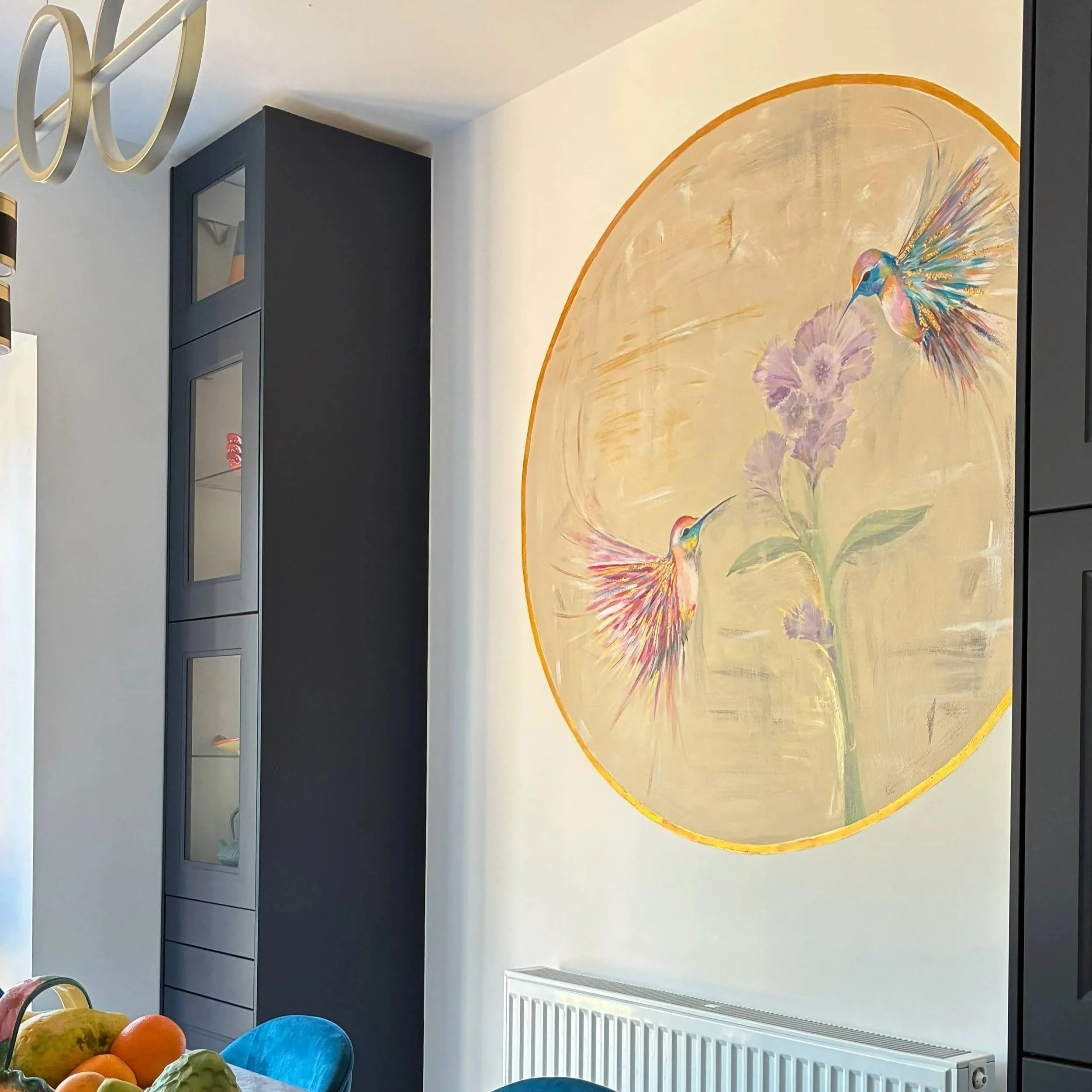 A colorful wall mural of a hummingbird hovering near purple flowers on a beige background, in a modern indoor space. A gray cabinet with glass doors is on the left and a white radiator is below the mural. A table with fruits, including oranges and ap