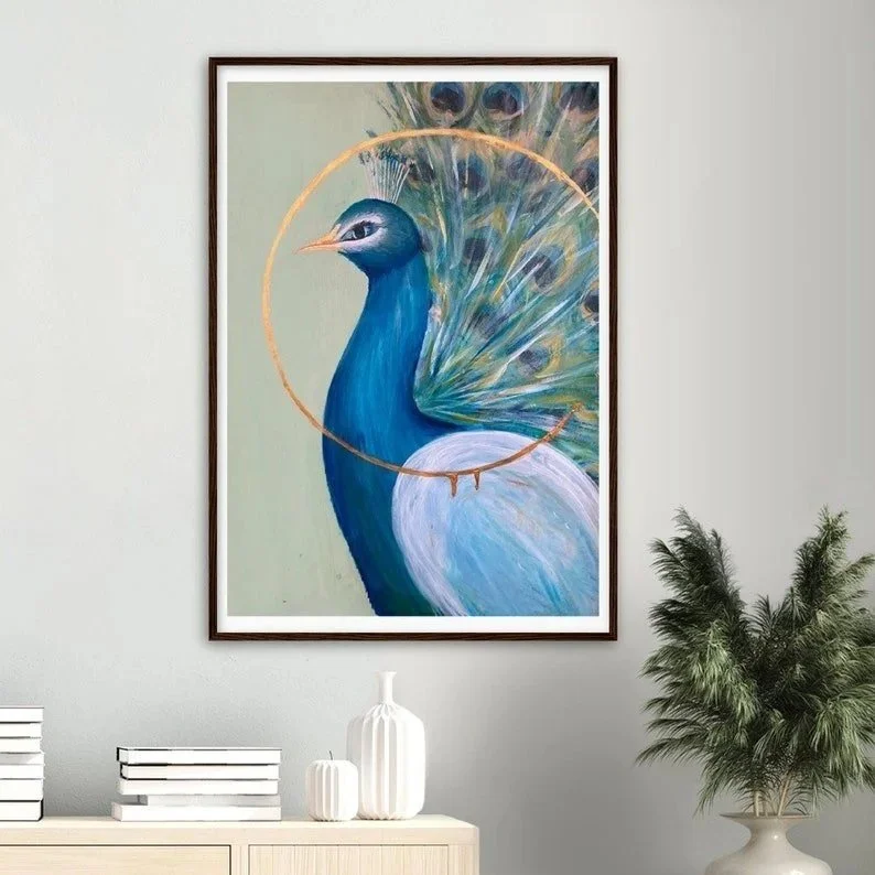 Painting of a blue peacock with its tail feathers fanned out, enclosed by a gold circle, hanging on a light gray wall with decor including books, vases, and a green plant.