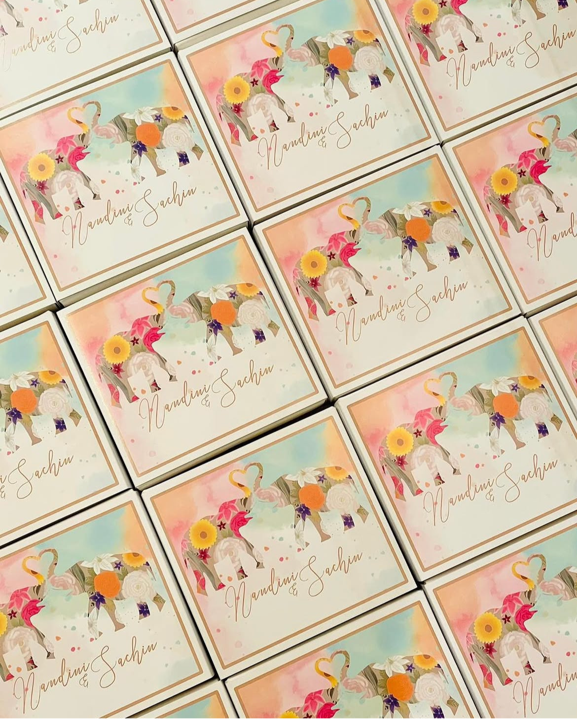 A collection of multiple colorful wedding favor boxes with a watercolor background, featuring an illustrated elephant with floral decorations, and the names 'Nandini & Sachin' written in elegant cursive.