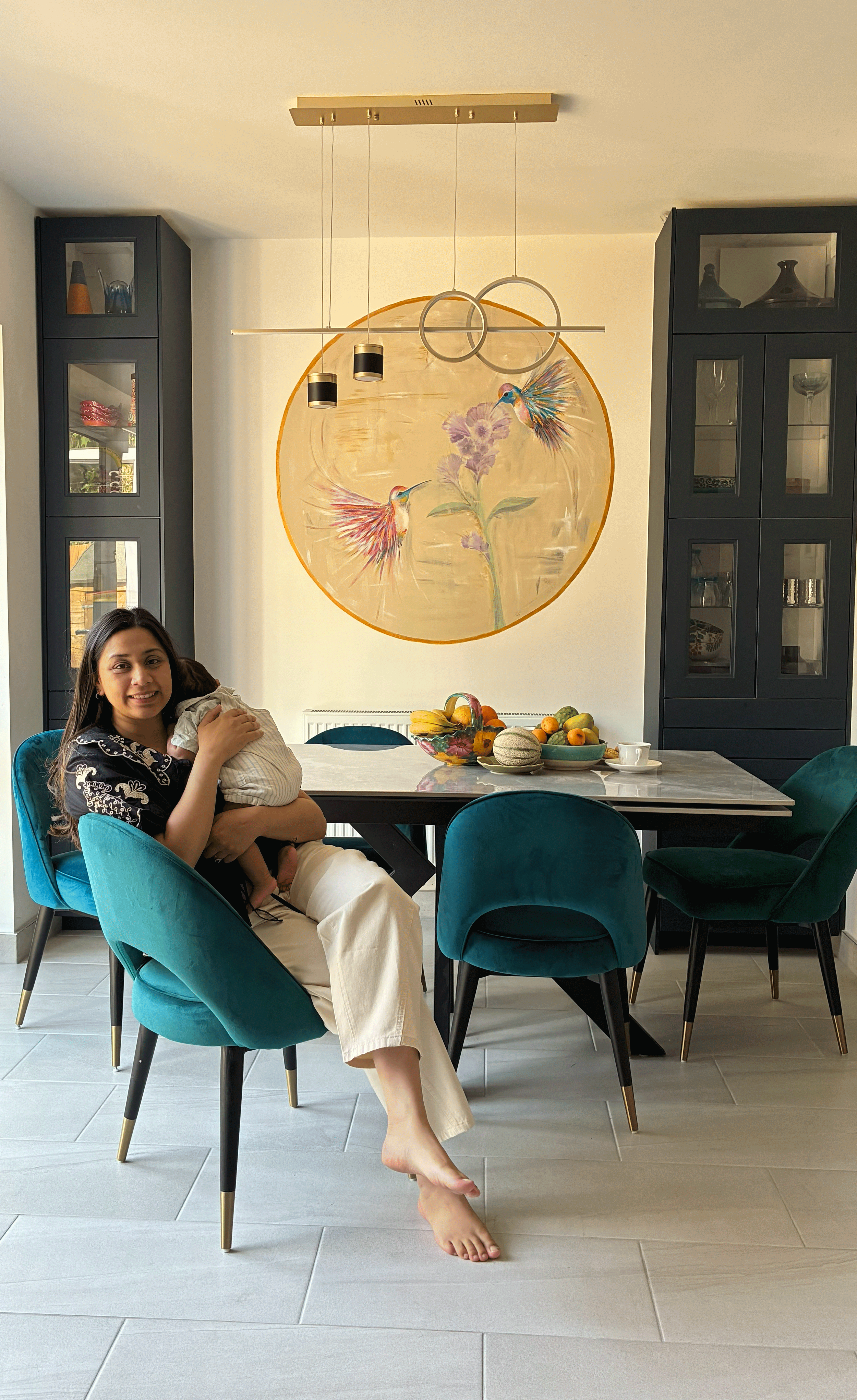 Woman sitting in a teal chair, hugging a child, in a dining room with a large circular wall art featuring hummingbirds and flowers, black cabinetry, and a table with fruit and a cup.