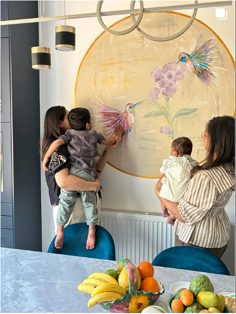 Two women with children look at a large wall painting of two colorful hummingbirds and purple flowers. One woman is holding a boy who is reaching toward the painting, while the other woman holds a girl. There is a table with bananas, apples, and oranges in the foreground.