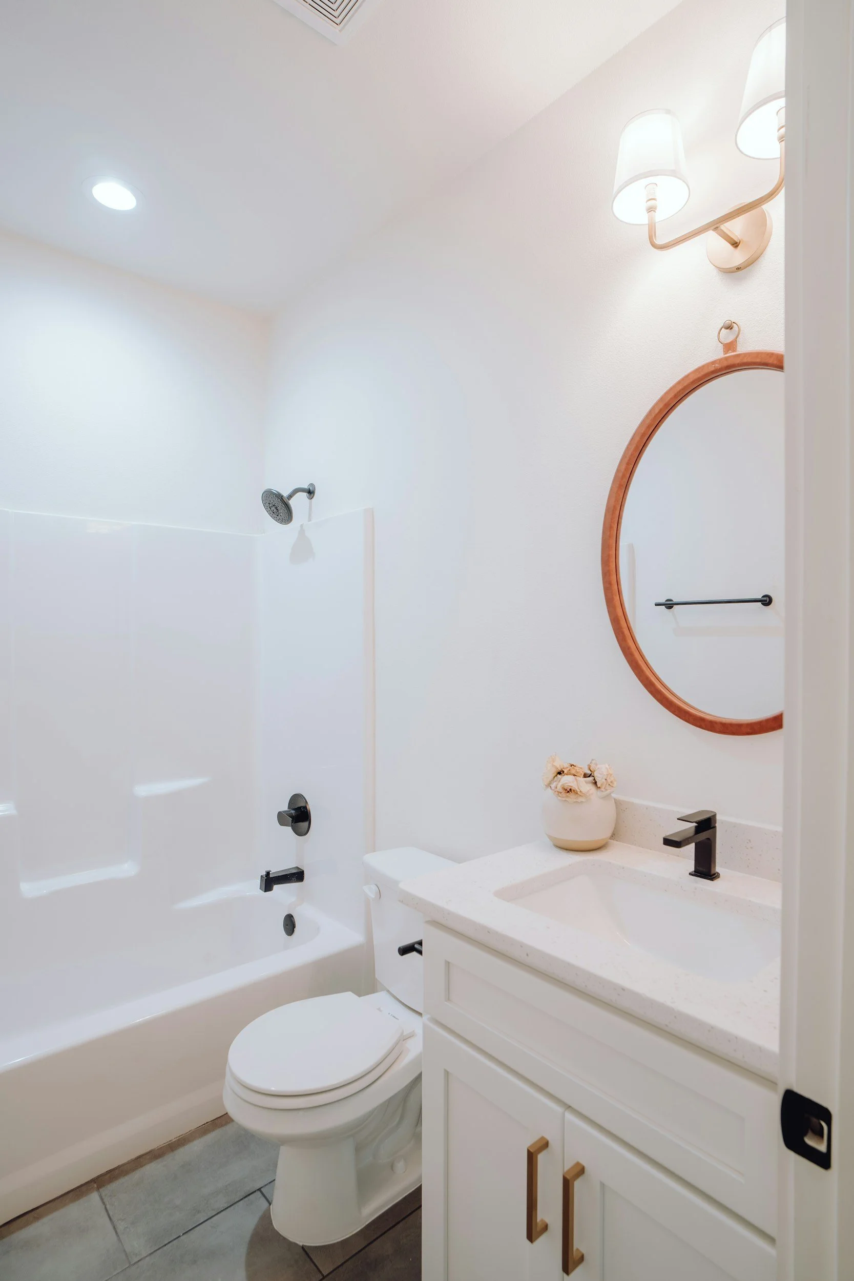 A modern bathroom featuring a bathtub with a showerhead, a toilet, a white vanity with a sink, a round mirror, a vase with flowers, and wall-mounted light fixtures.