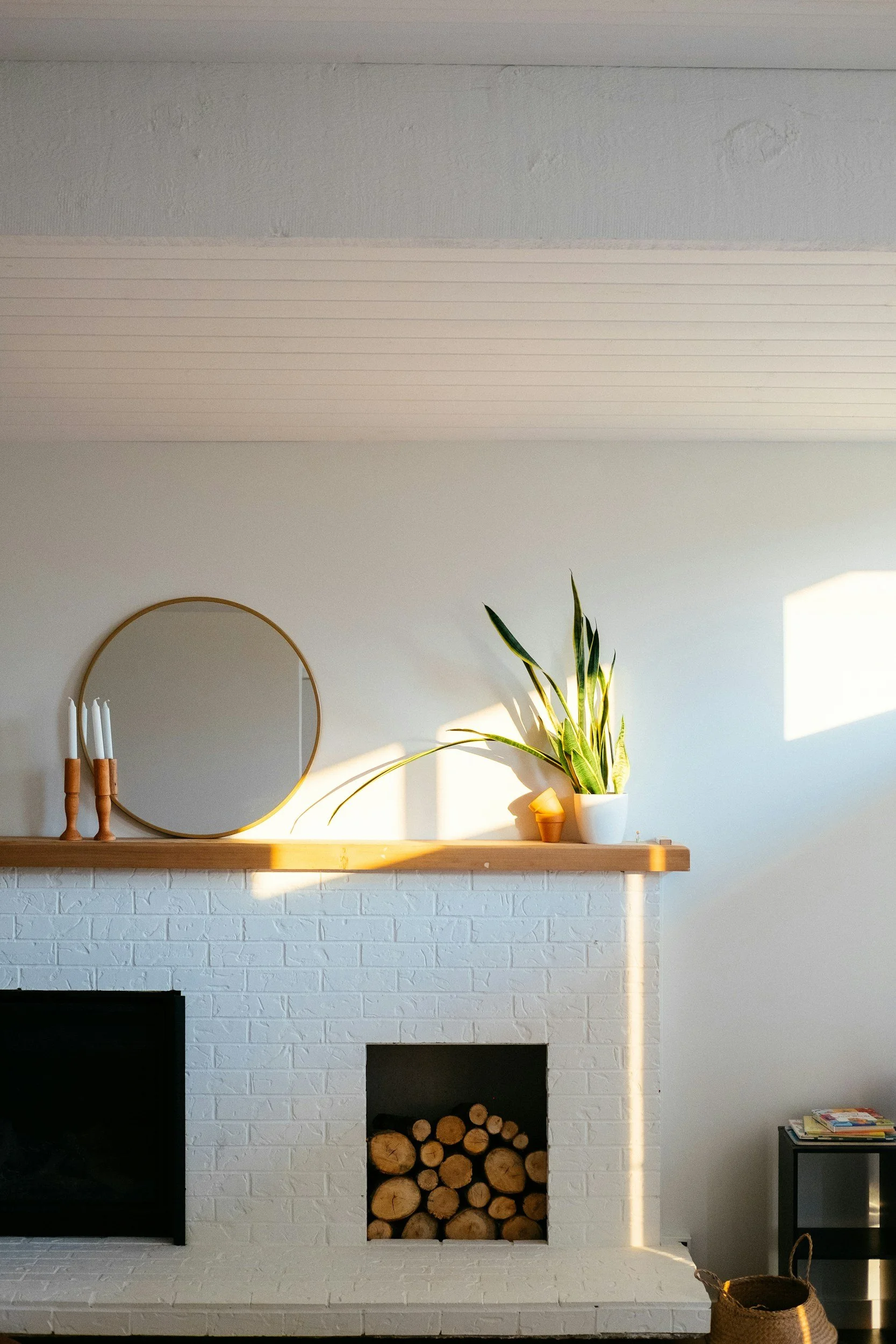 Living room with white brick fireplace, open logs, and wooden mantel decorated with a round mirror, white candles in wooden holders, a potted plant, a small terracotta pot, and a stack of books, illuminated by sunlight.