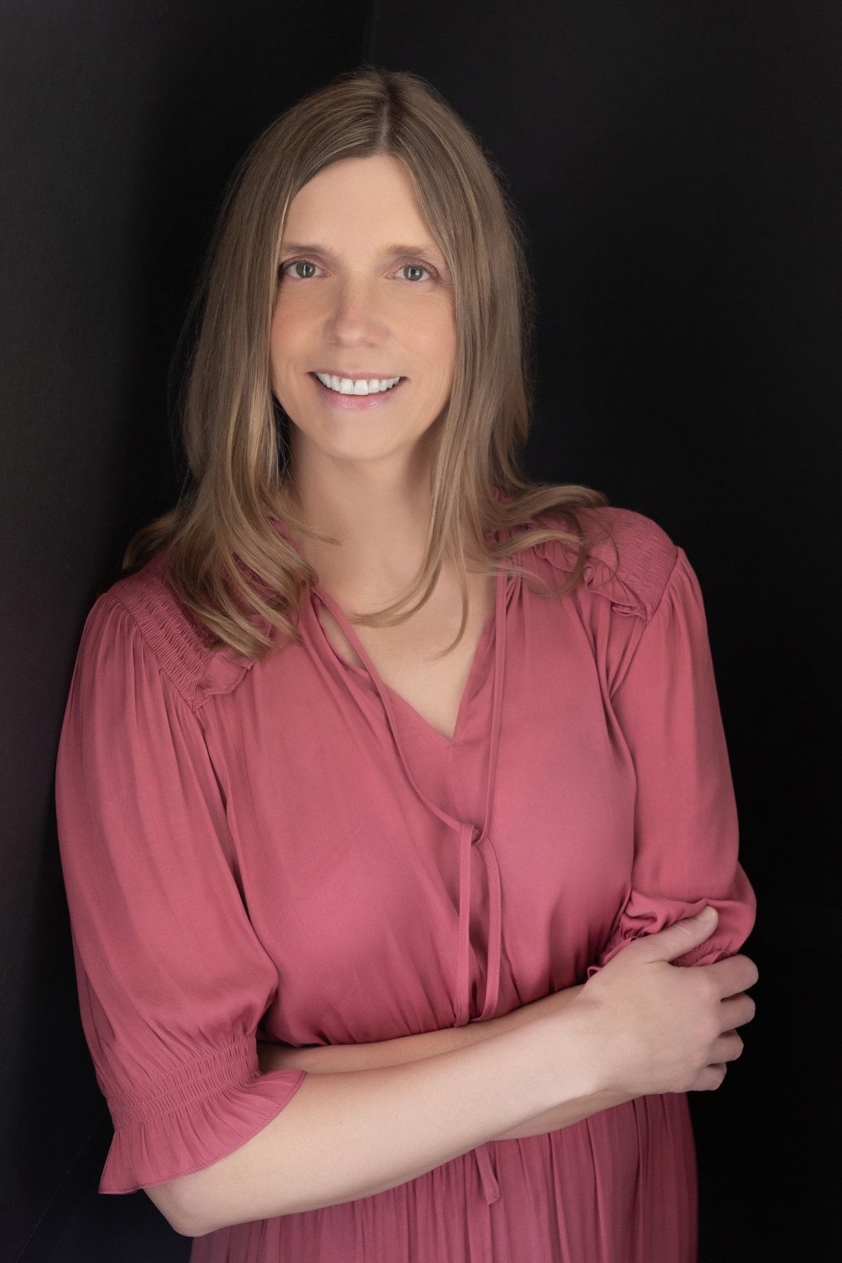 A woman with shoulder-length light brown hair wearing a pink dress, smiling, with her arms crossed against a black background.