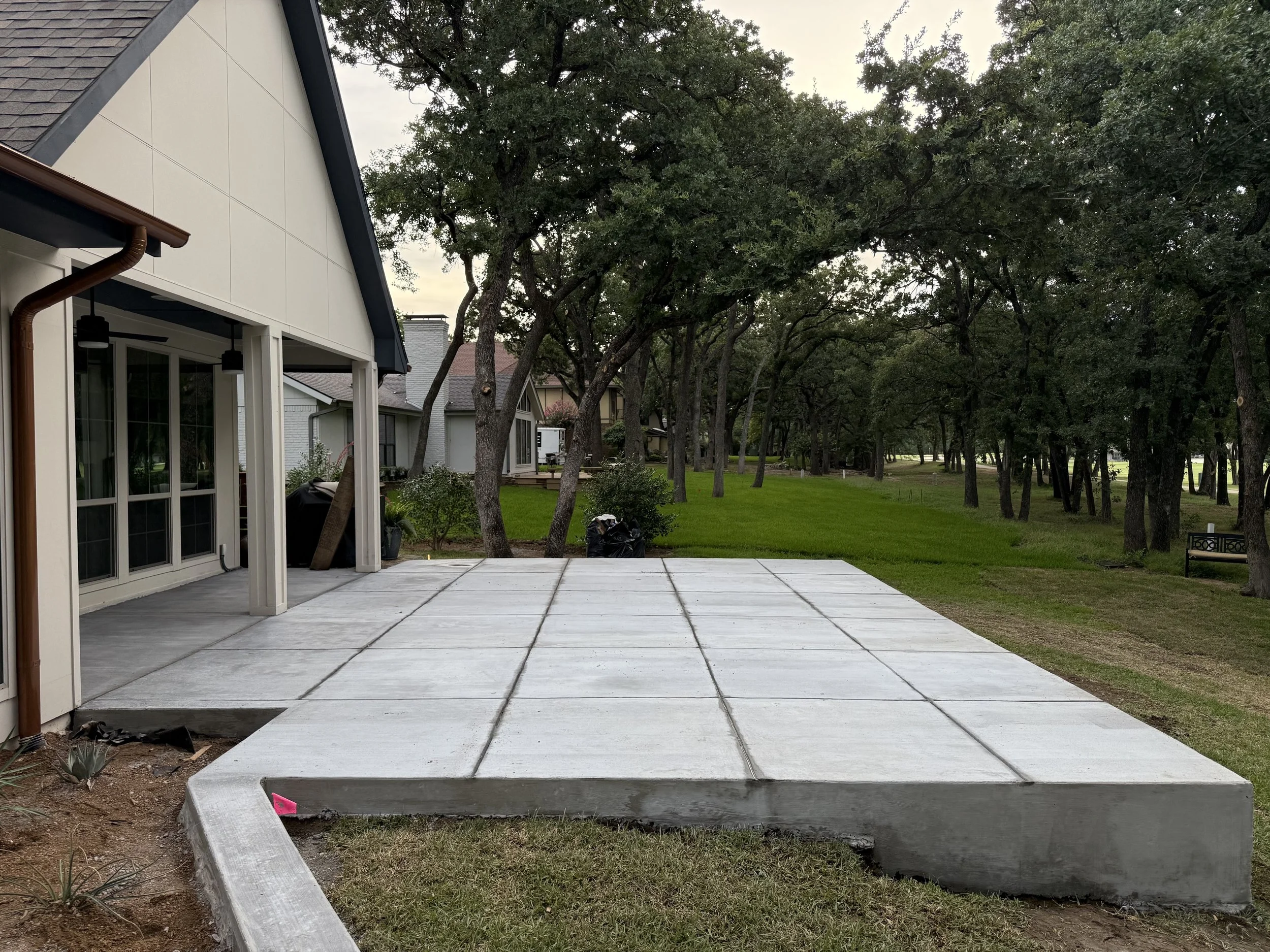 A newly poured concrete patio in a suburban backyard with neighboring houses and green trees in the background.