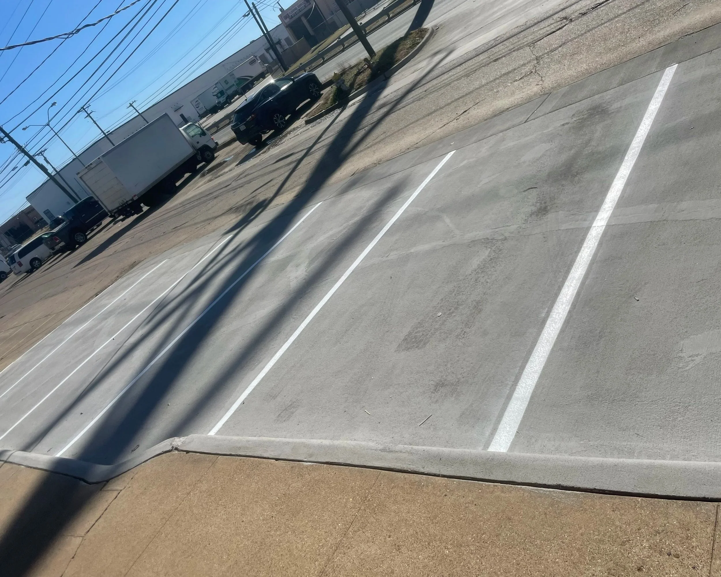 A parking lot with several cars and trucks parked along a sidewalk under a clear blue sky. Power lines cast shadows across the pavement.