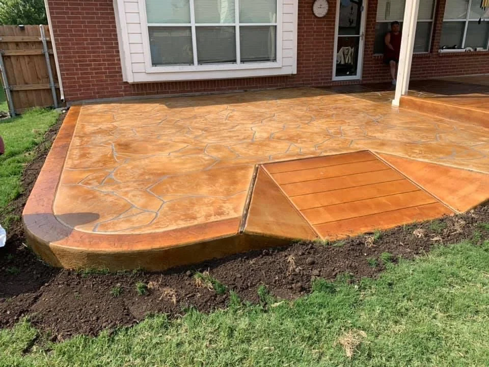 Newly installed stained concrete patio with a wood plank pattern on one section, in backyard next to house with window and door, and a person standing in the doorway.
