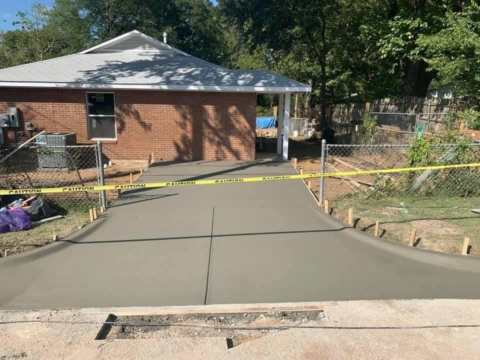 Newly poured concrete driveway in front of a brick house with a trailer and backyard fencing, marked off with caution tape