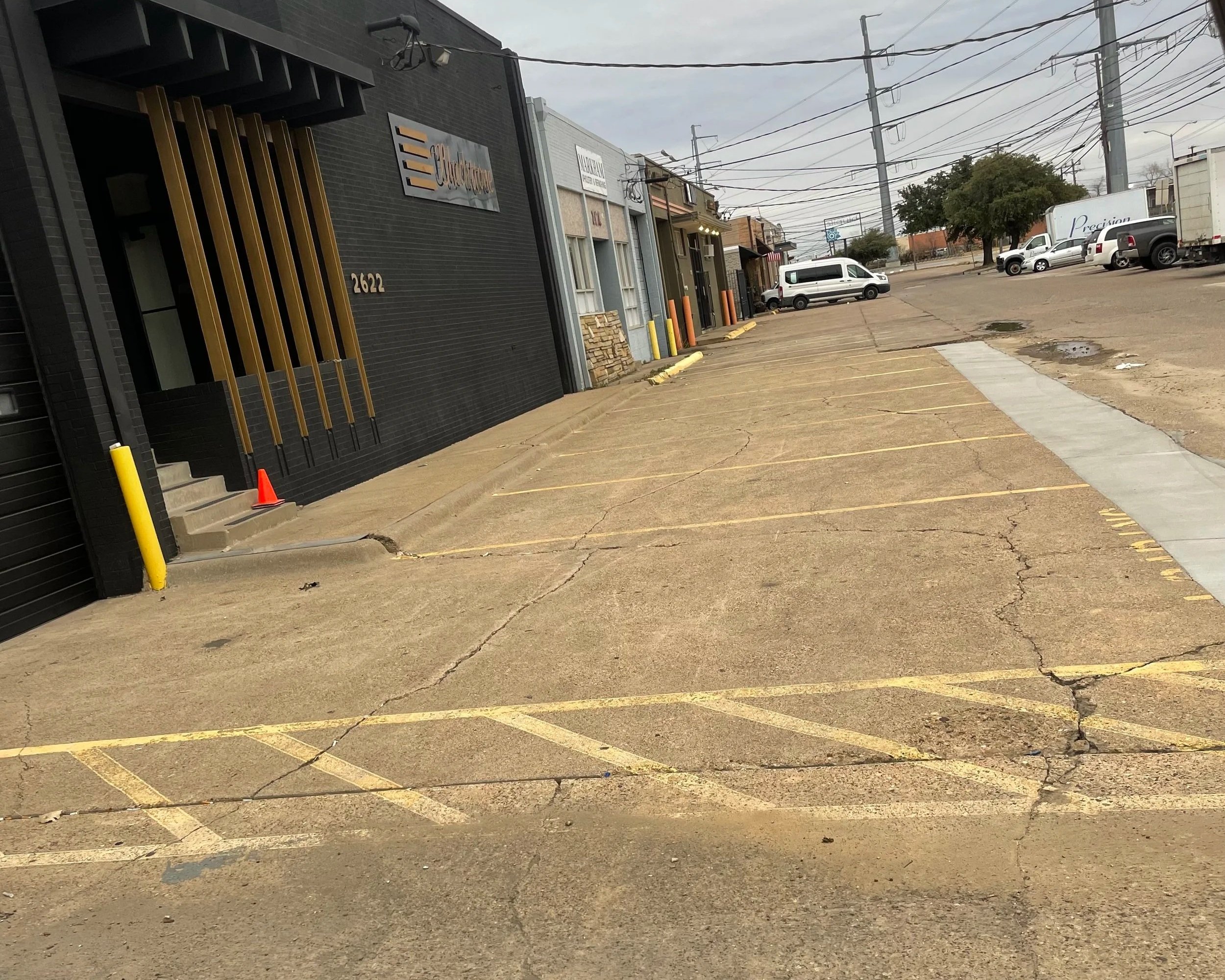 Paved parking lot with yellow painted parking lines, some cracks, and puddles. Commercial buildings in the background with signs and parked vehicles, including a white van and trucks, under a cloudy sky.
