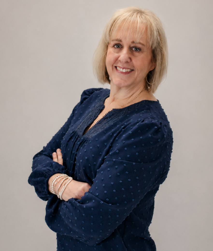 A blonde woman in a navy blue dress with polka dots, smiling with arms crossed, wearing jewelry, in front of a plain gray background.
