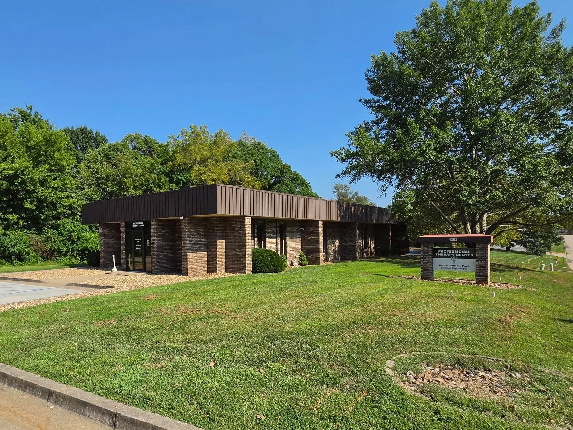 Building of a professional therapy center with brick exterior and a sign in front displaying the name, 'Professional Therapy Center,' and the name 'Ann M. Pollock, PsyD,' surrounded by green lawn and trees under a clear blue sky.