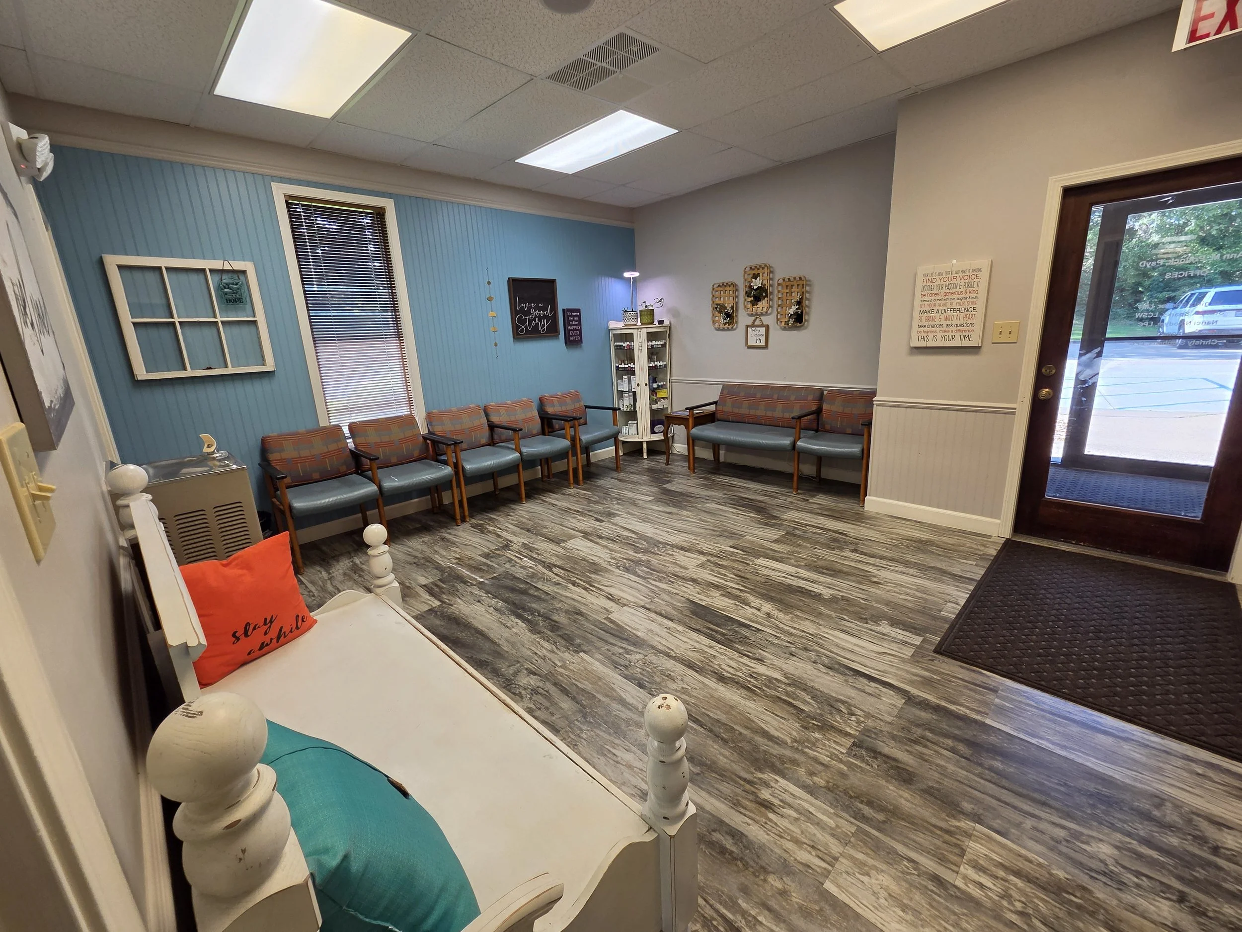 Waiting room with blue wall, multiple chairs, a small cabinet with supplies, wall decorations, and a door with a window showing parked cars outside.