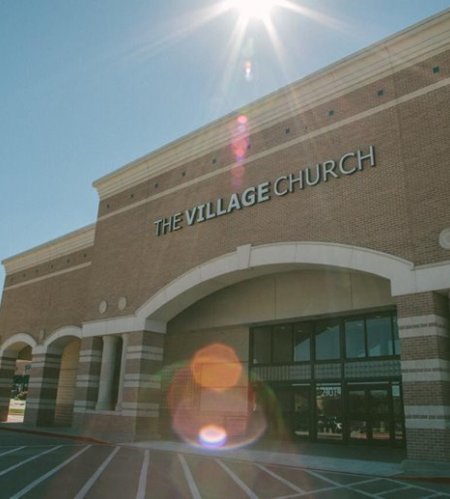 The exterior of a church building named 'The Village Church' under a bright sunny sky with sun glare and lens flares.