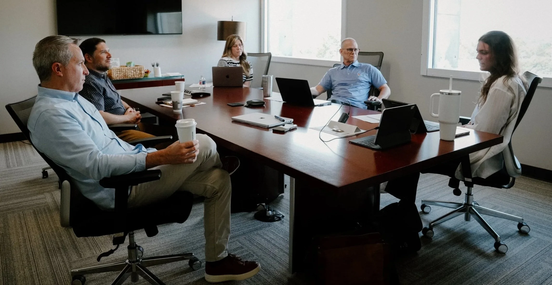 Five people sitting around a conference table in a business meeting room, with laptops, cups, and documents.