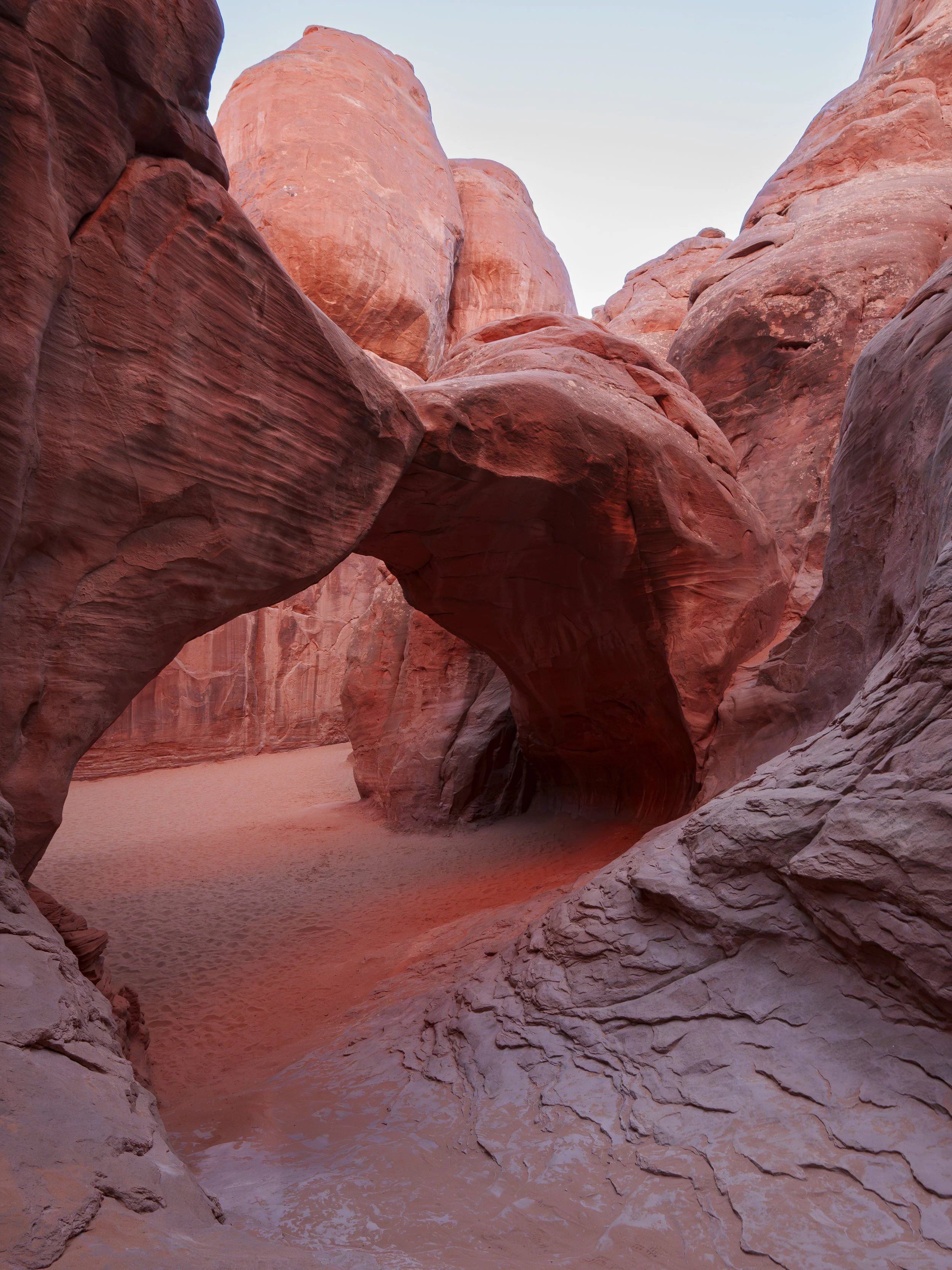 Photo of a slot canyon with smooth red rock formations and a sandy floor, with a natural rock arch in the middle of the scene.