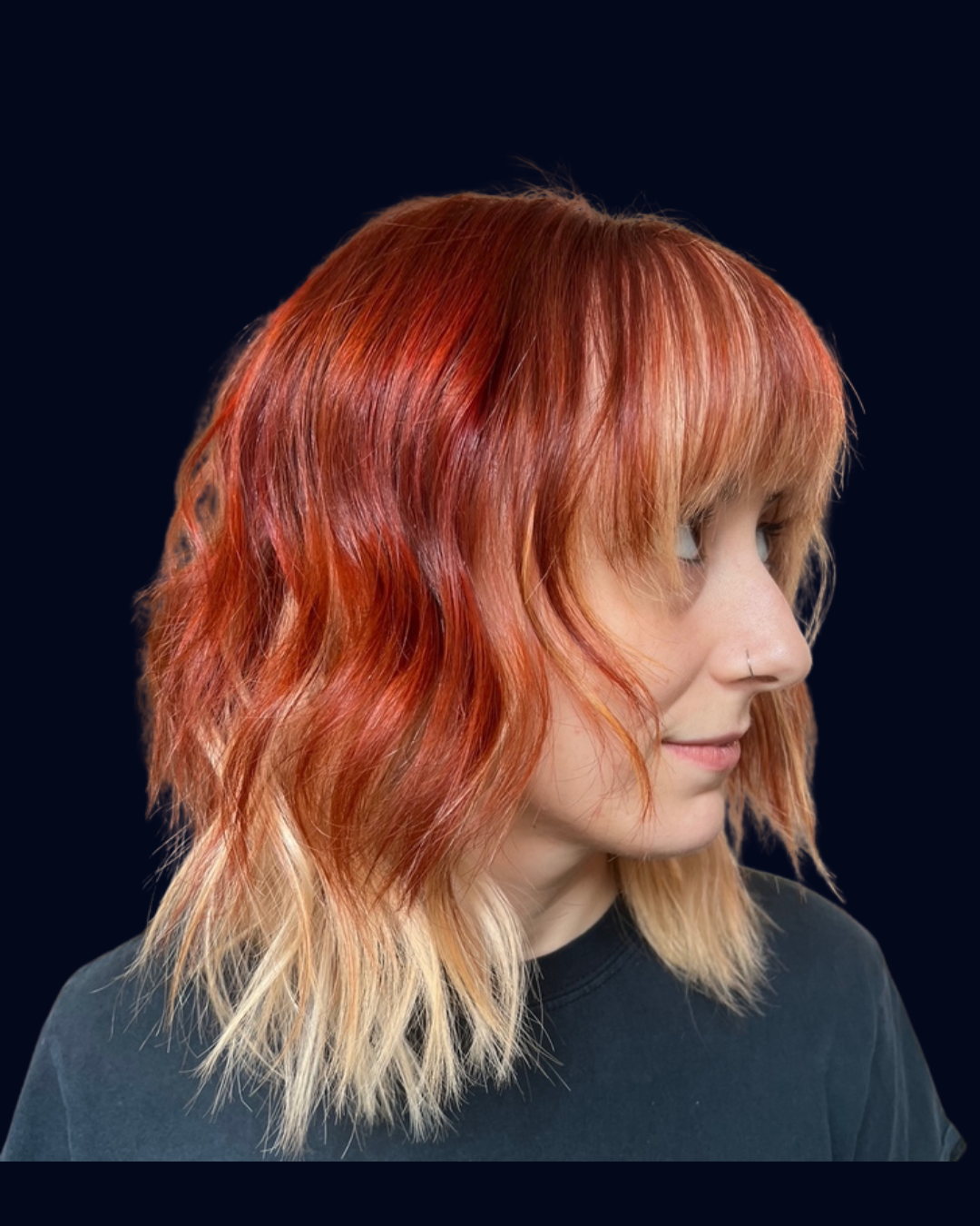 Woman with shoulder-length wavy red hair and blonde ends, wearing a nose ring, facing right, against a dark background.
