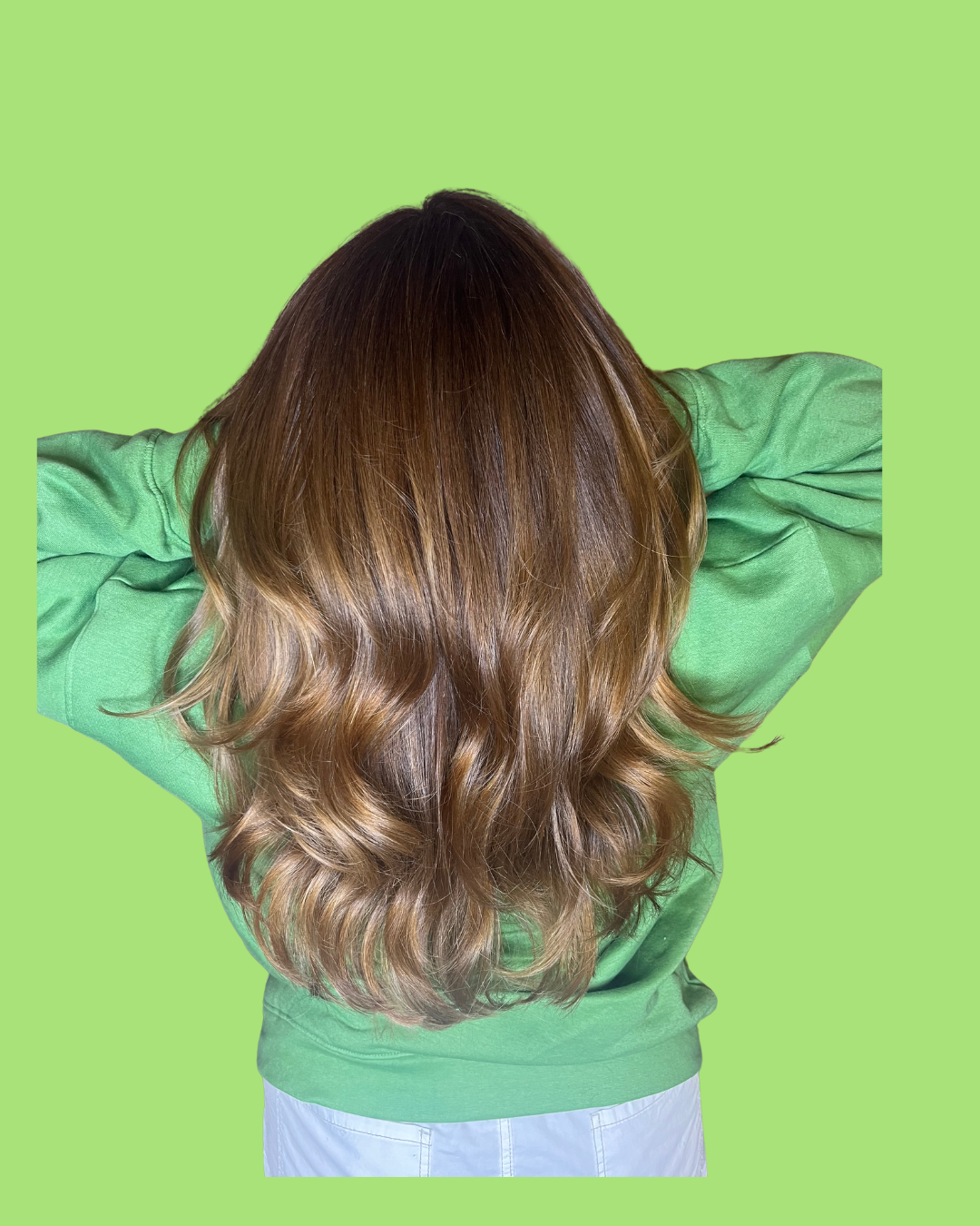 Back view of a person with long, wavy, brown hair wearing a green sweatshirt and light-colored pants, with their hands behind their head against a green background.