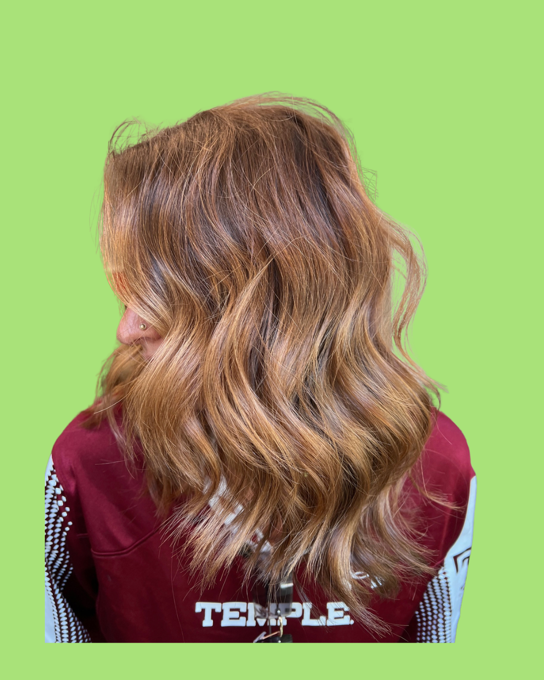 Side view of a woman with shoulder-length, wavy, auburn hair, wearing a maroon jacket with white text on the back, against a light green background.