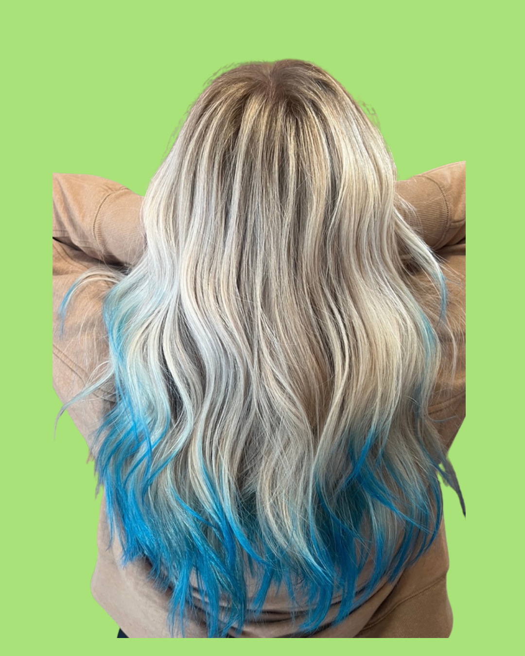 Back view of a woman with long, wavy blonde hair with blue tips, wearing a tan jacket, against a green background.