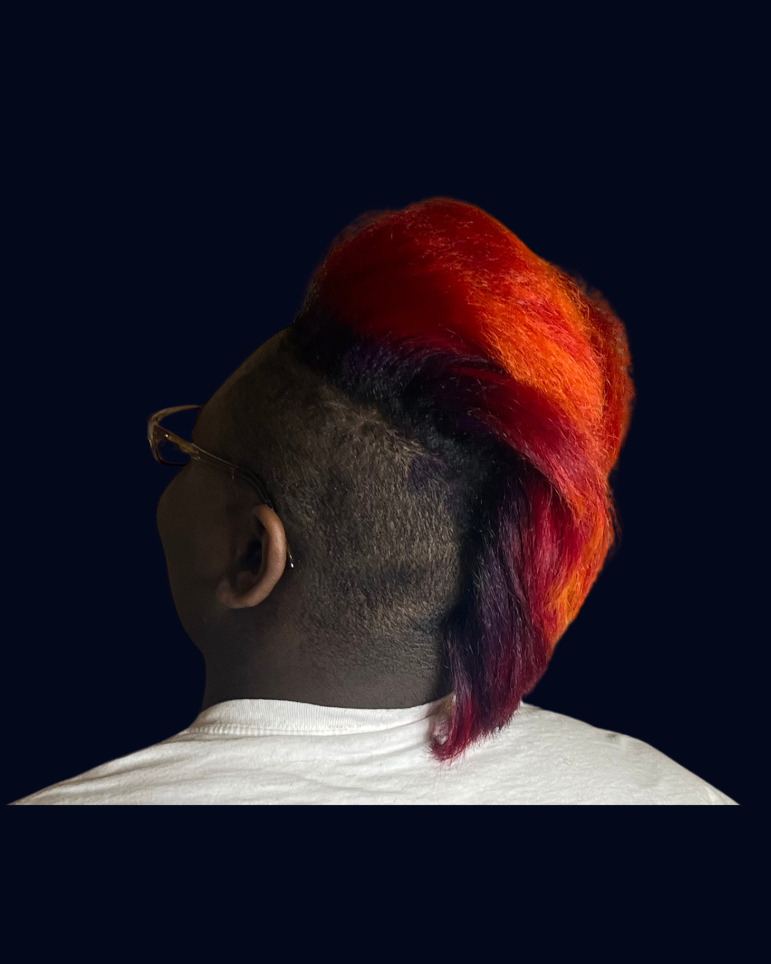 Back view of a person with a dark skin tone and freshly dyed vibrant red and orange mohawk hairstyle, wearing glasses and a white shirt, against a dark background.