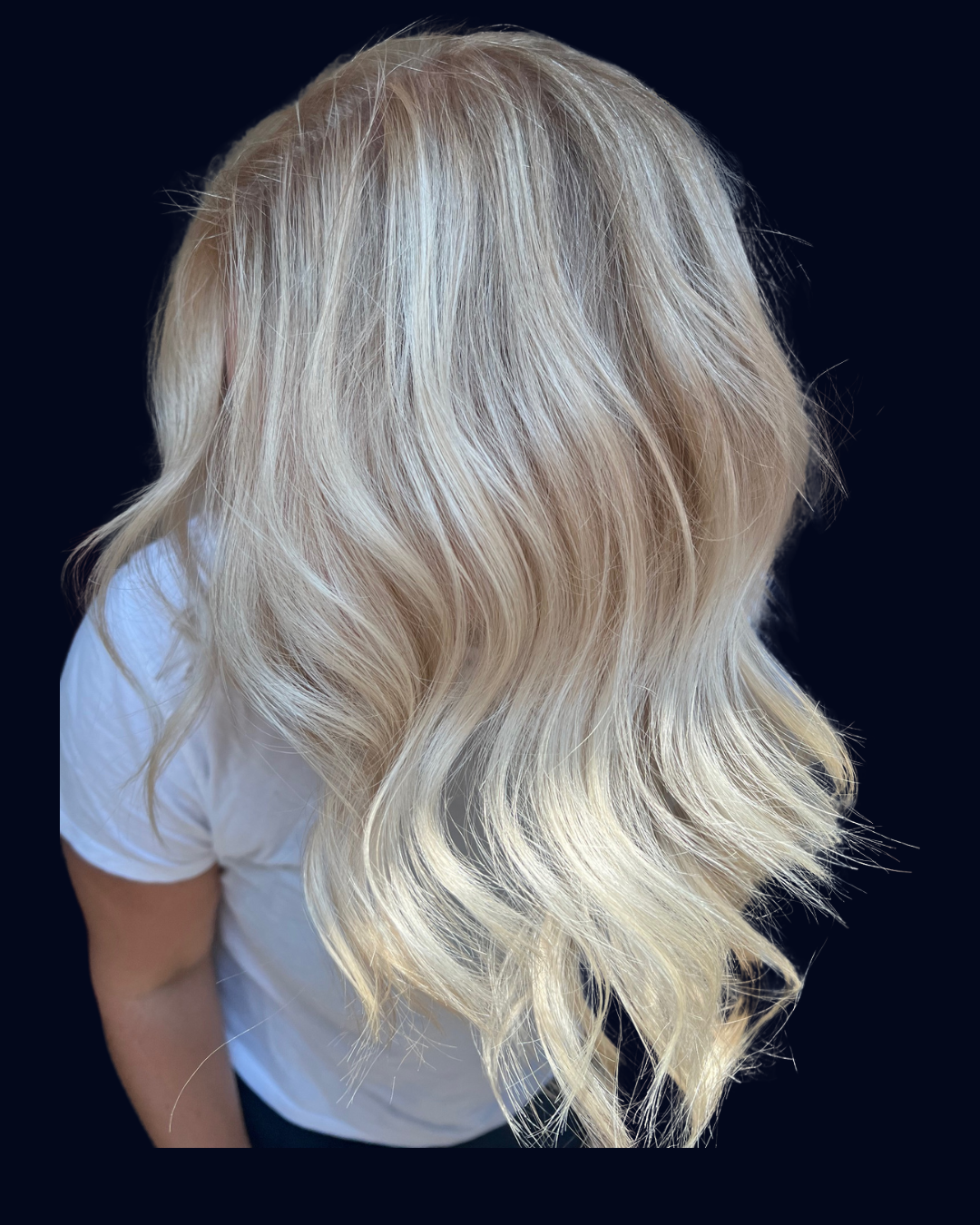 Blonde, wavy hair viewed from the back, wearing a white shirt against a dark background.