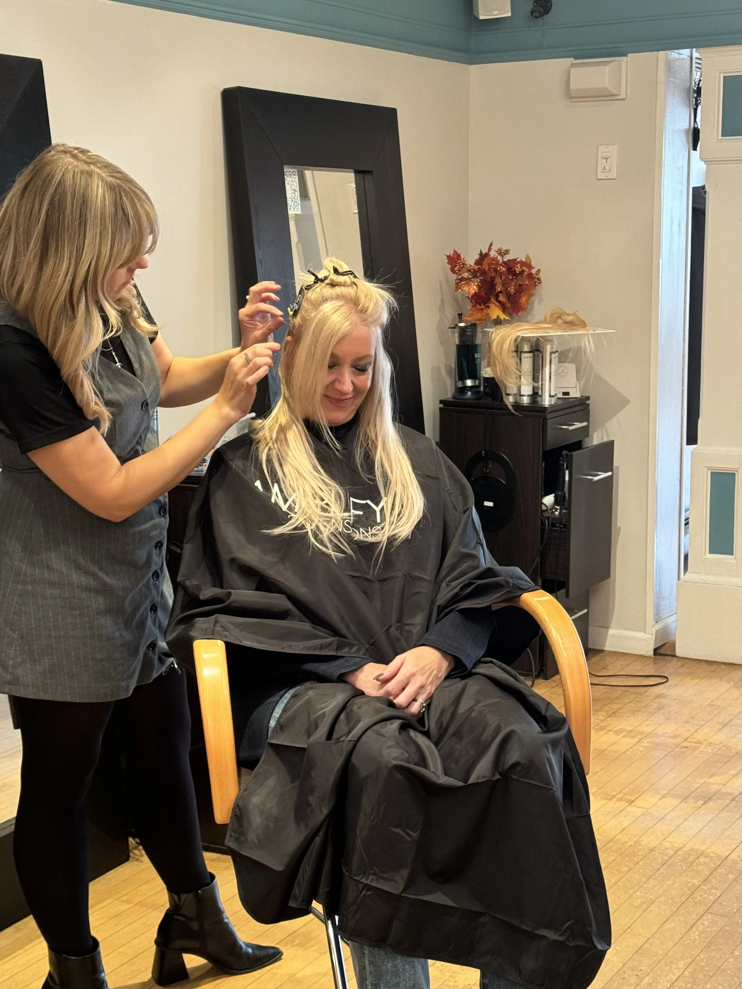 A woman with long blonde hair sitting in a salon chair, receiving a hair treatment, with a stylist working on her hair. The salon has light-colored walls decorated with autumn leaves and hair products on a nearby shelf.