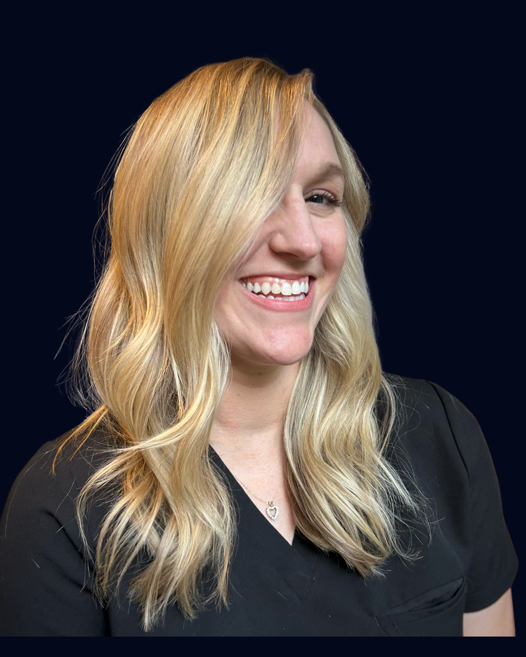 A smiling woman with long blond hair, wearing a black top and a necklace with a heart pendant, against a dark background.