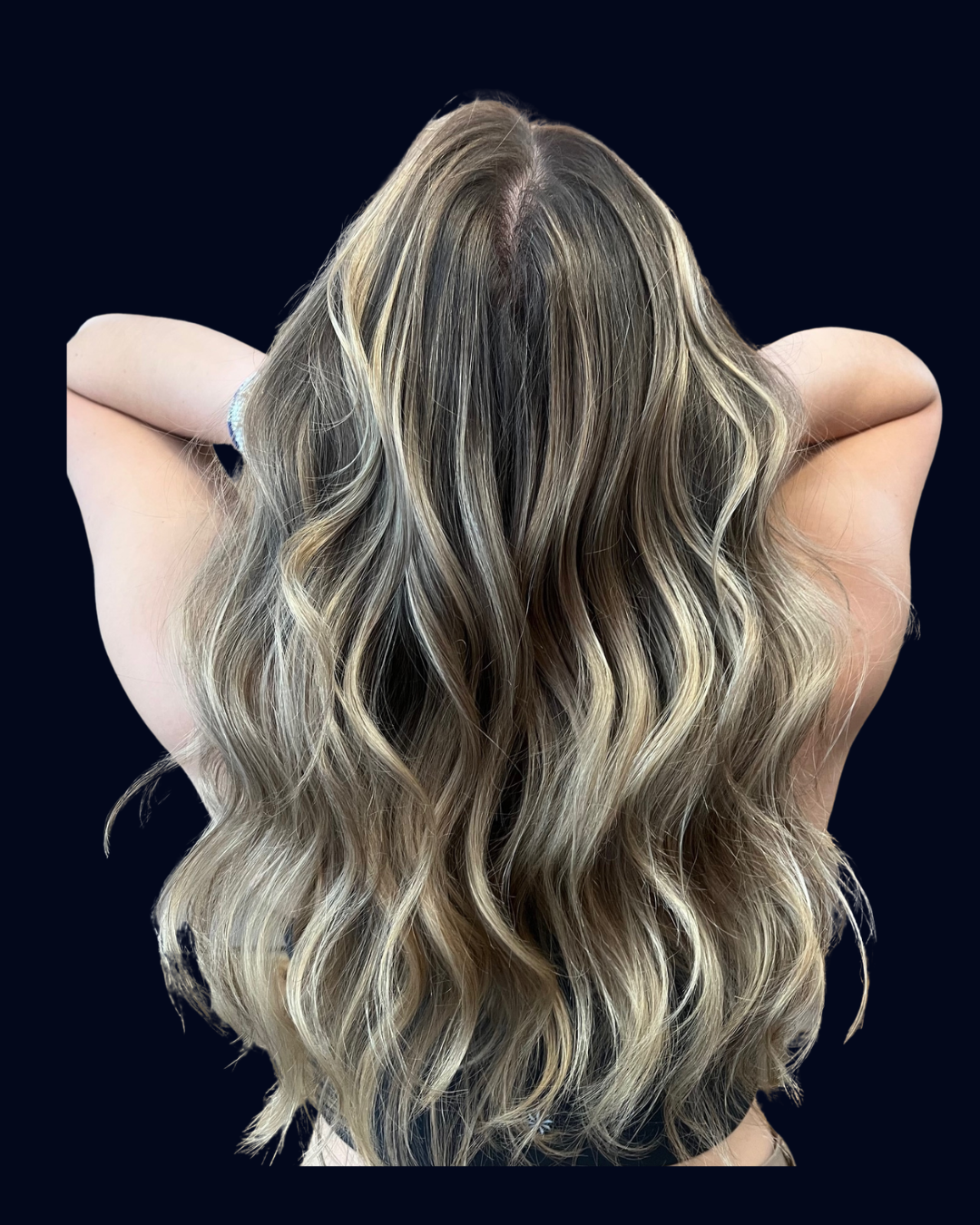 Back view of a person with long, wavy blonde hair, arms raised behind their head, against a black background.