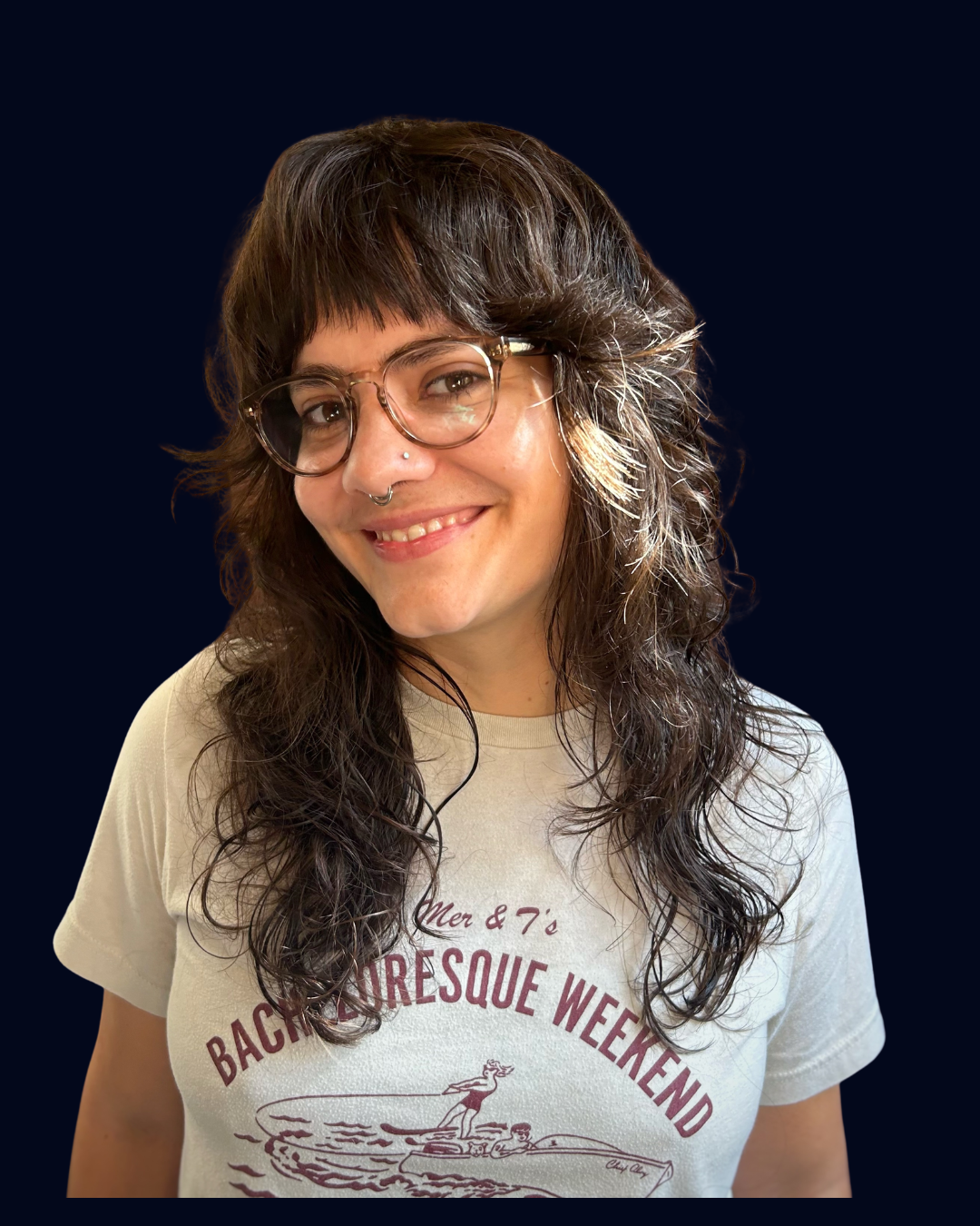 Smiling woman with glasses and curly hair, wearing a T-shirt with a retro-style design, posing against a dark background.